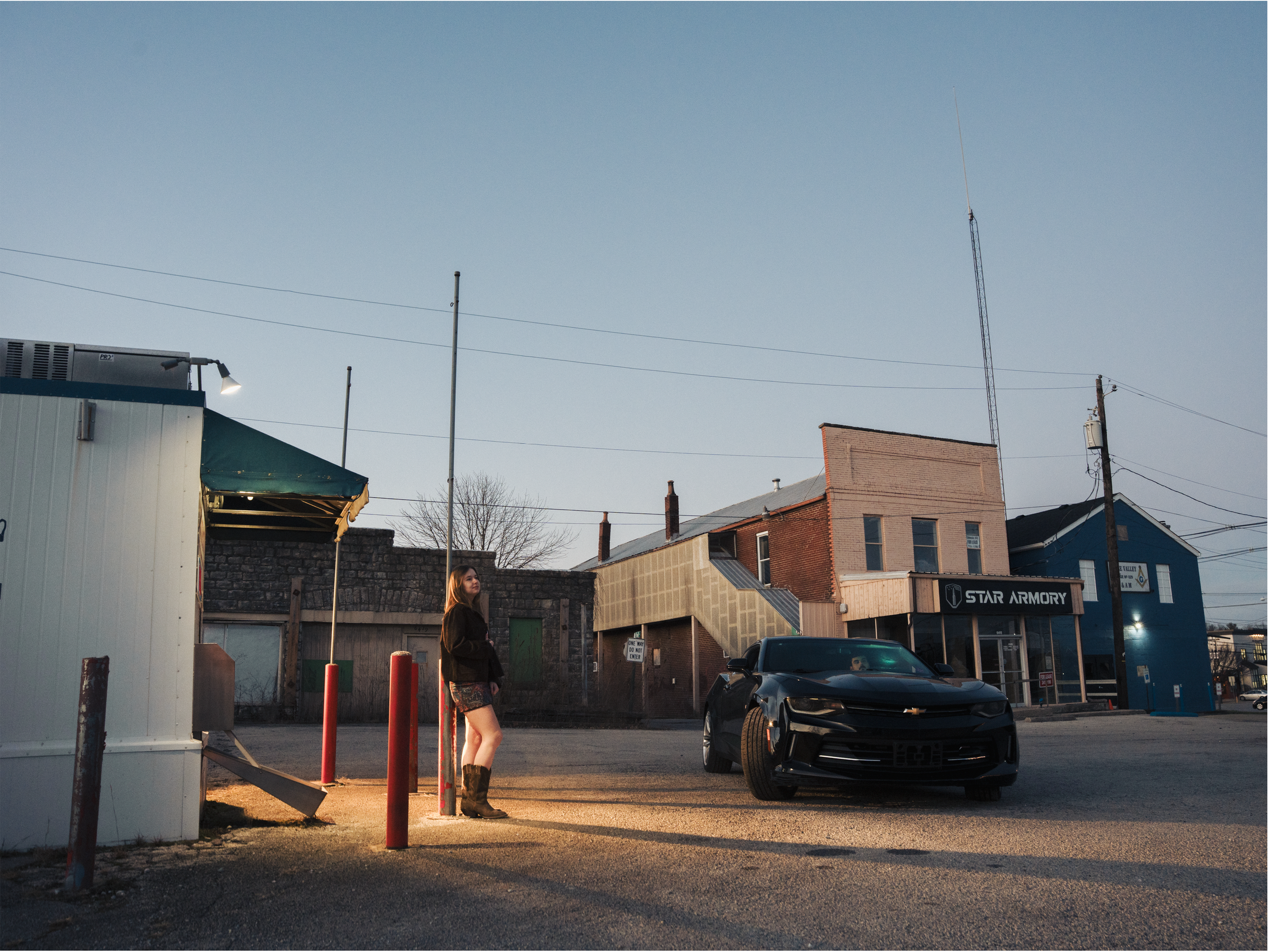 A young woman with long hair wearing a black jacket, patterned shorts, and boots, standing near red posts and a white building, with a black Chevrolet Camaro parked in front, in an urban area during late afternoon or early evening with clear sky.
