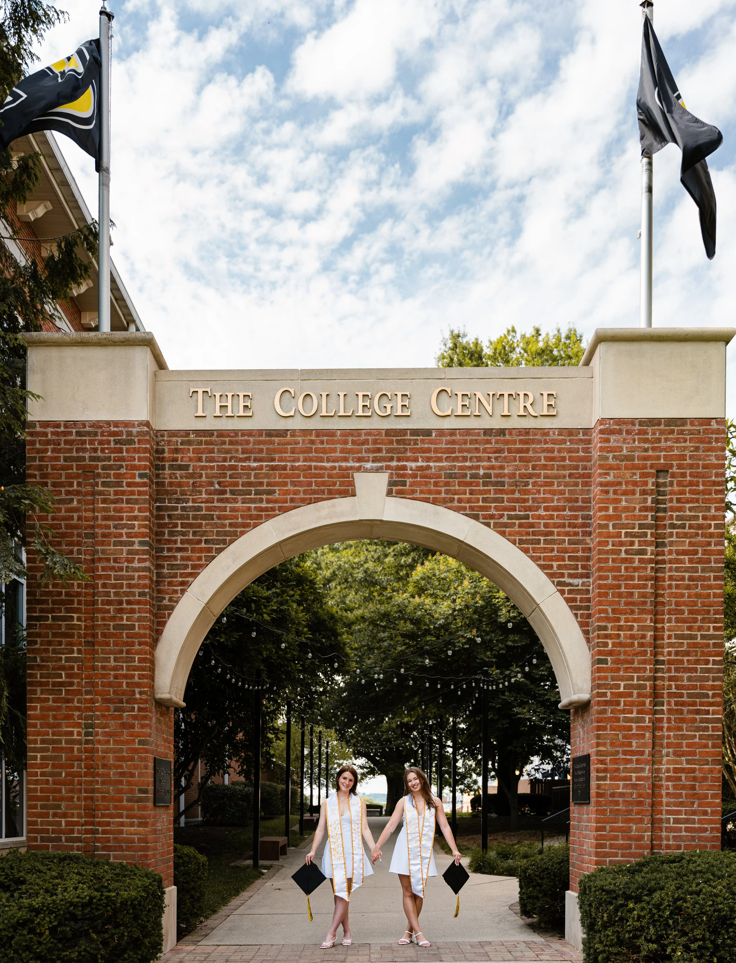 Two women in graduation gowns and caps holding hands below a brick archway at The College Centre, with trees and a blue sky with clouds in the background.