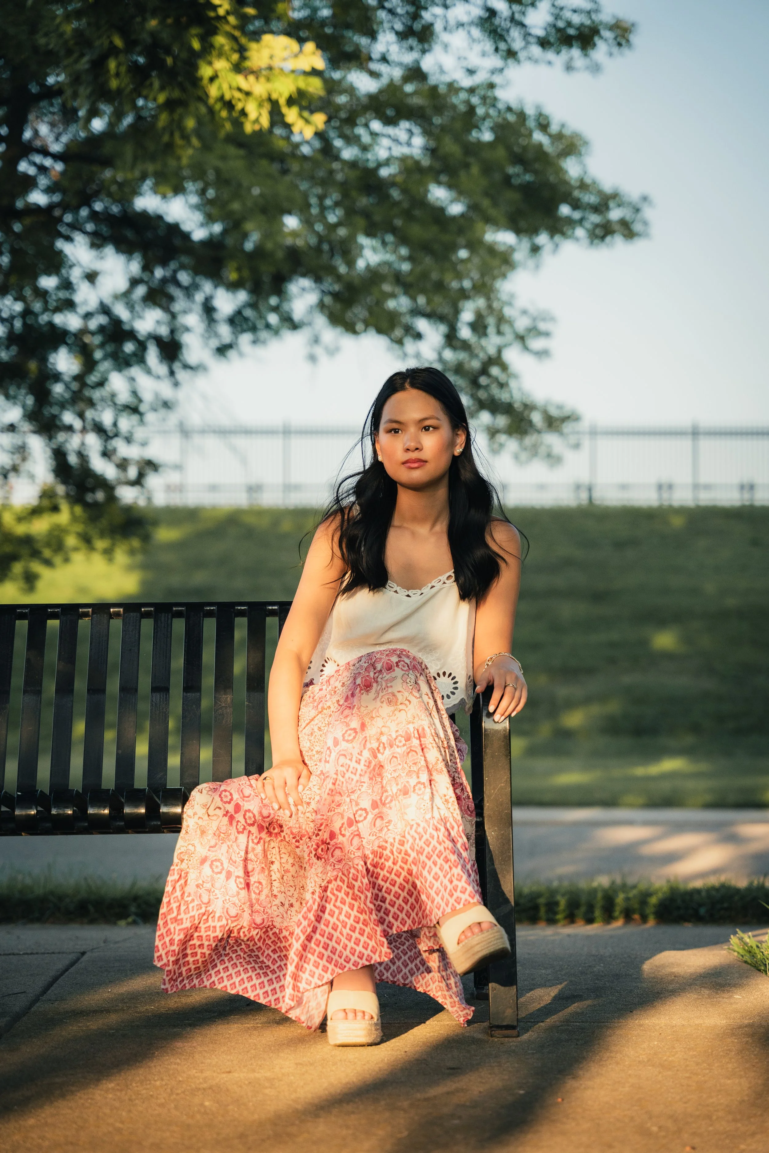 A young woman with long dark hair sitting on a black metal bench outdoors. She is wearing a white sleeveless top and a long pink skirt with a pattern. She has a neutral expression and is looking slightly to her left. There are green trees in the back