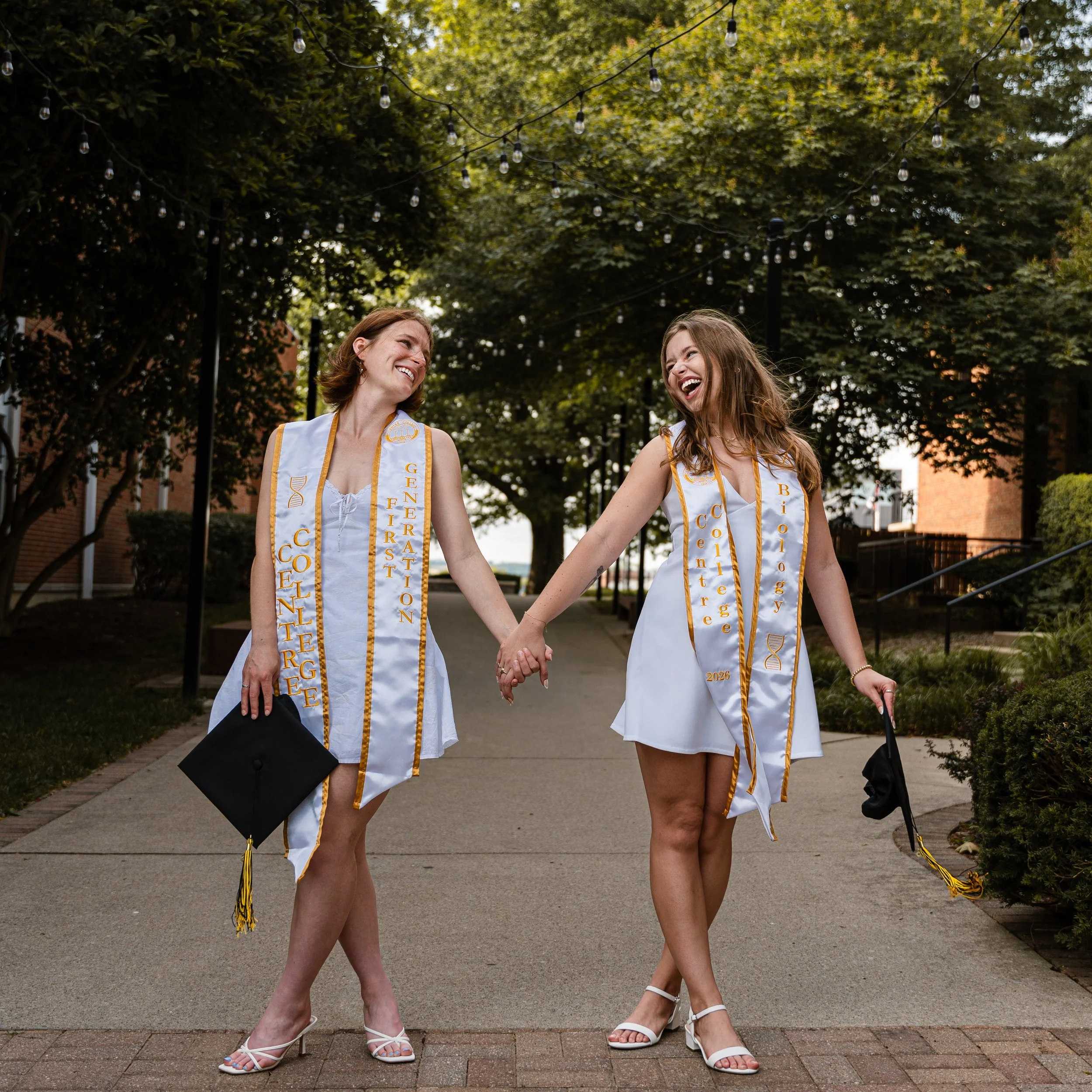 Two women in white dresses with yellow and black honor cords, holding hands and smiling at each other outdoors under string lights and trees.
