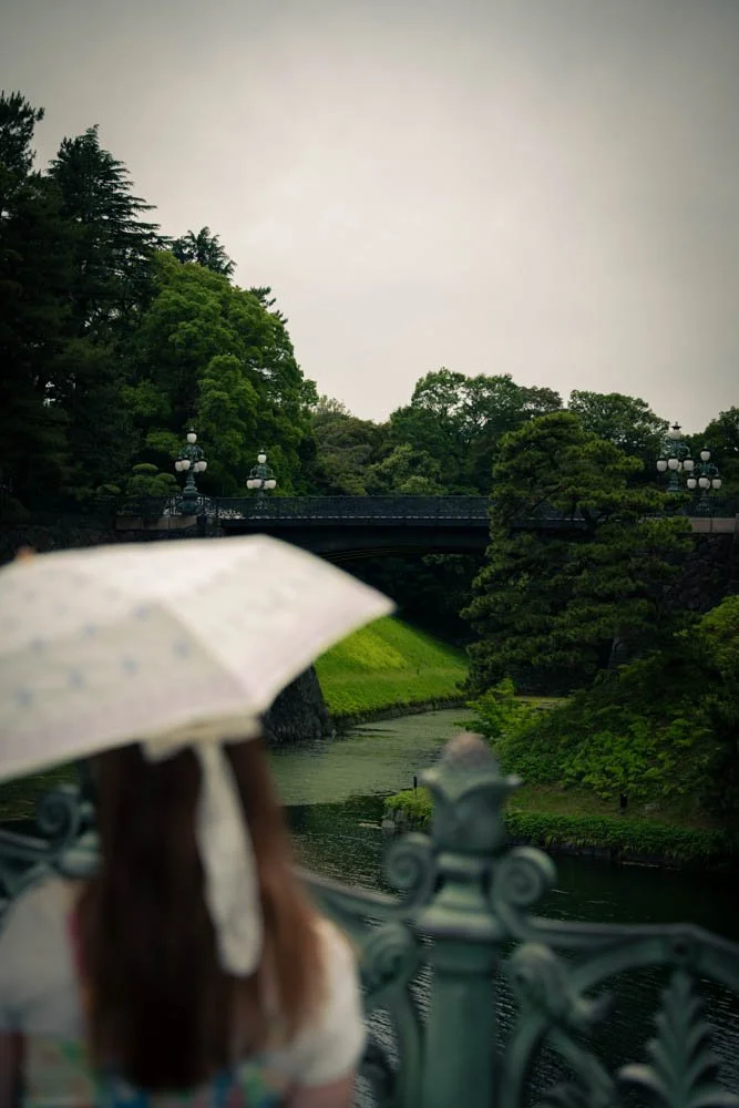 A person with long hair holding a white umbrella near a river, with greenery and trees in the background.