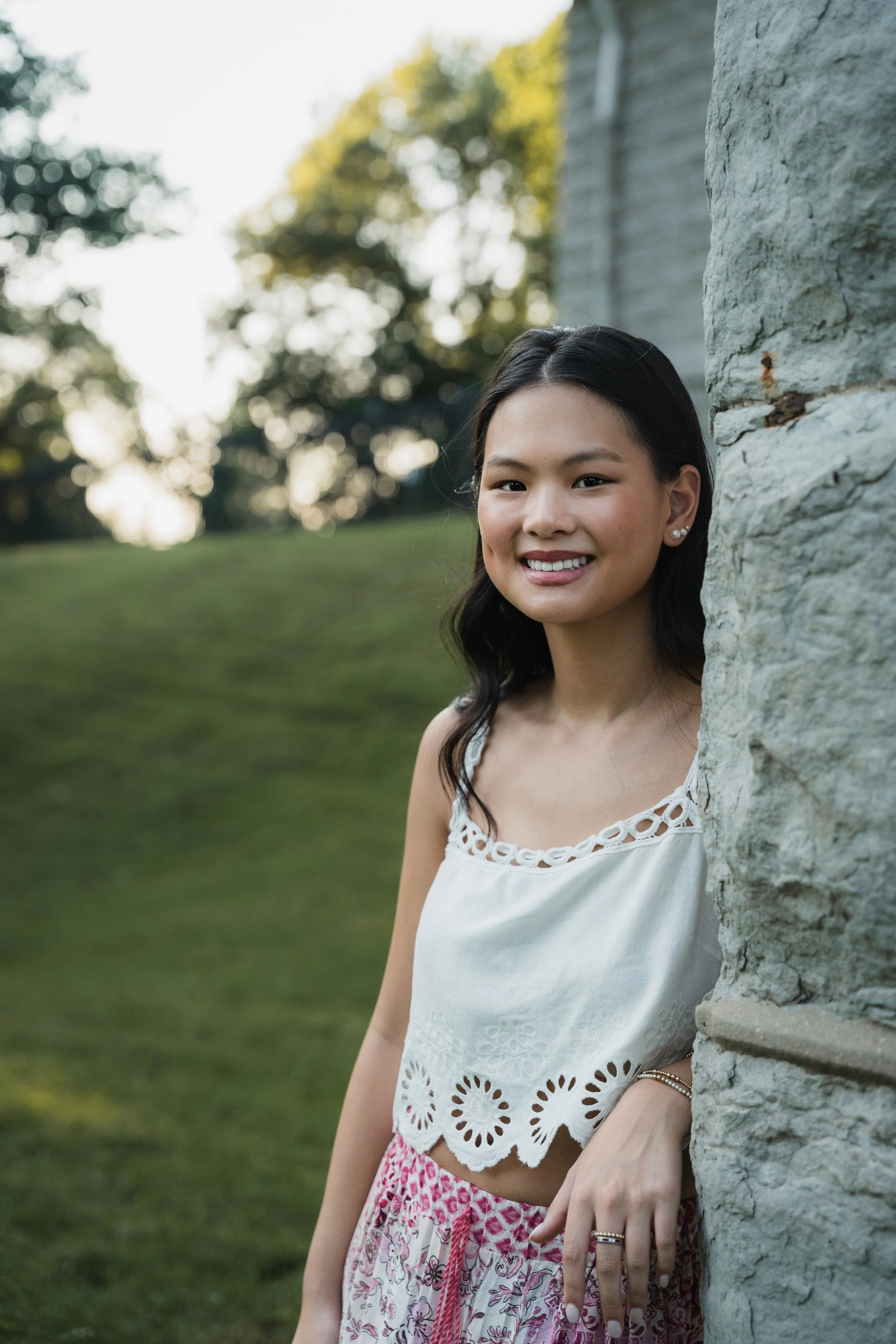 A young woman with dark hair, smiling, stands outdoors near a stone wall with trees and grass in the background during the daytime.