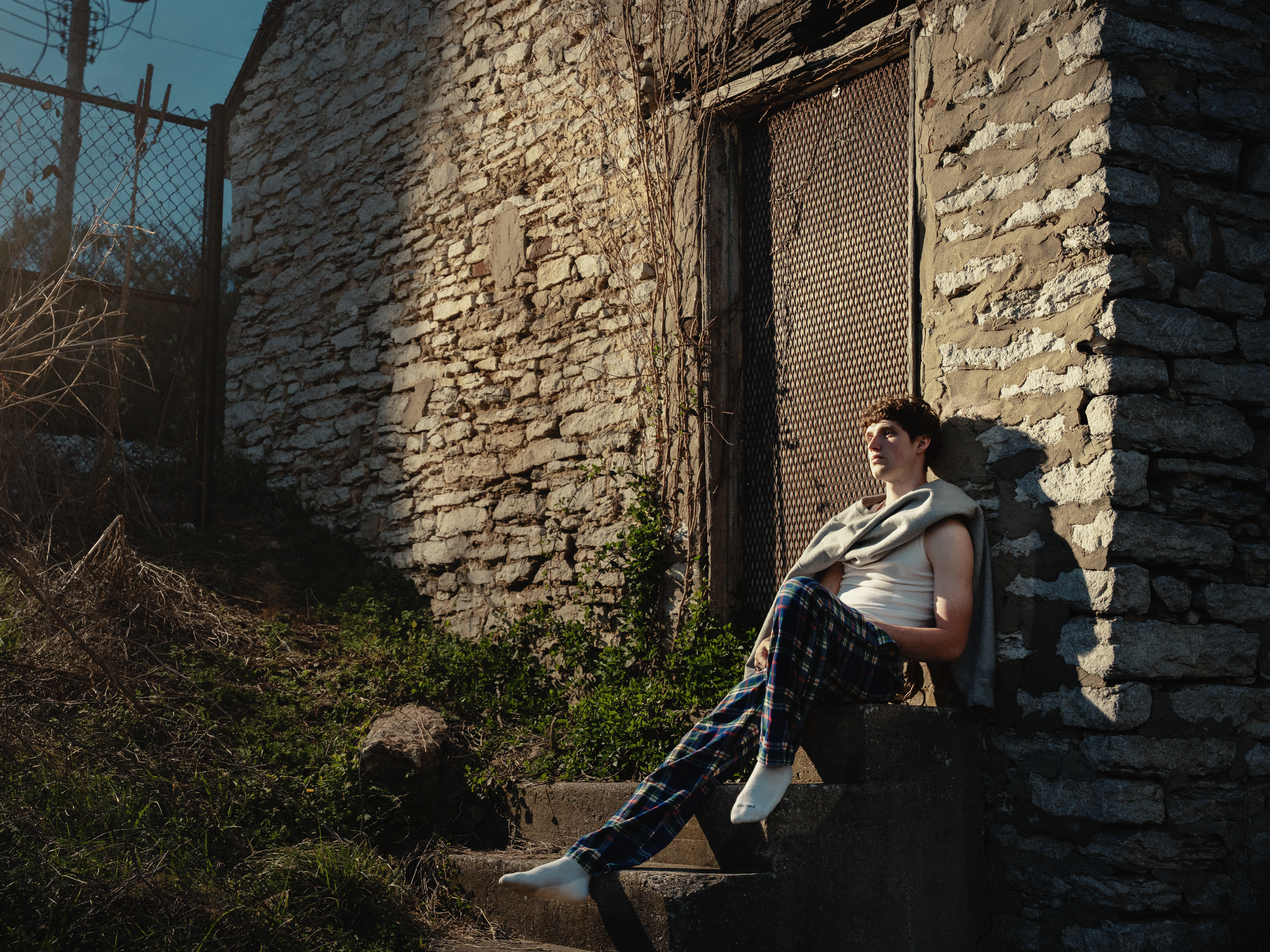 A young man with curly hair wearing a white hoodie and plaid pants sits on a concrete step next to a weathered stone wall with vines growing on it. There is a chain-link fence and electrical poles in the background, with some plants and shrubbery at the base of the wall. The lighting suggests it is late afternoon or early evening.