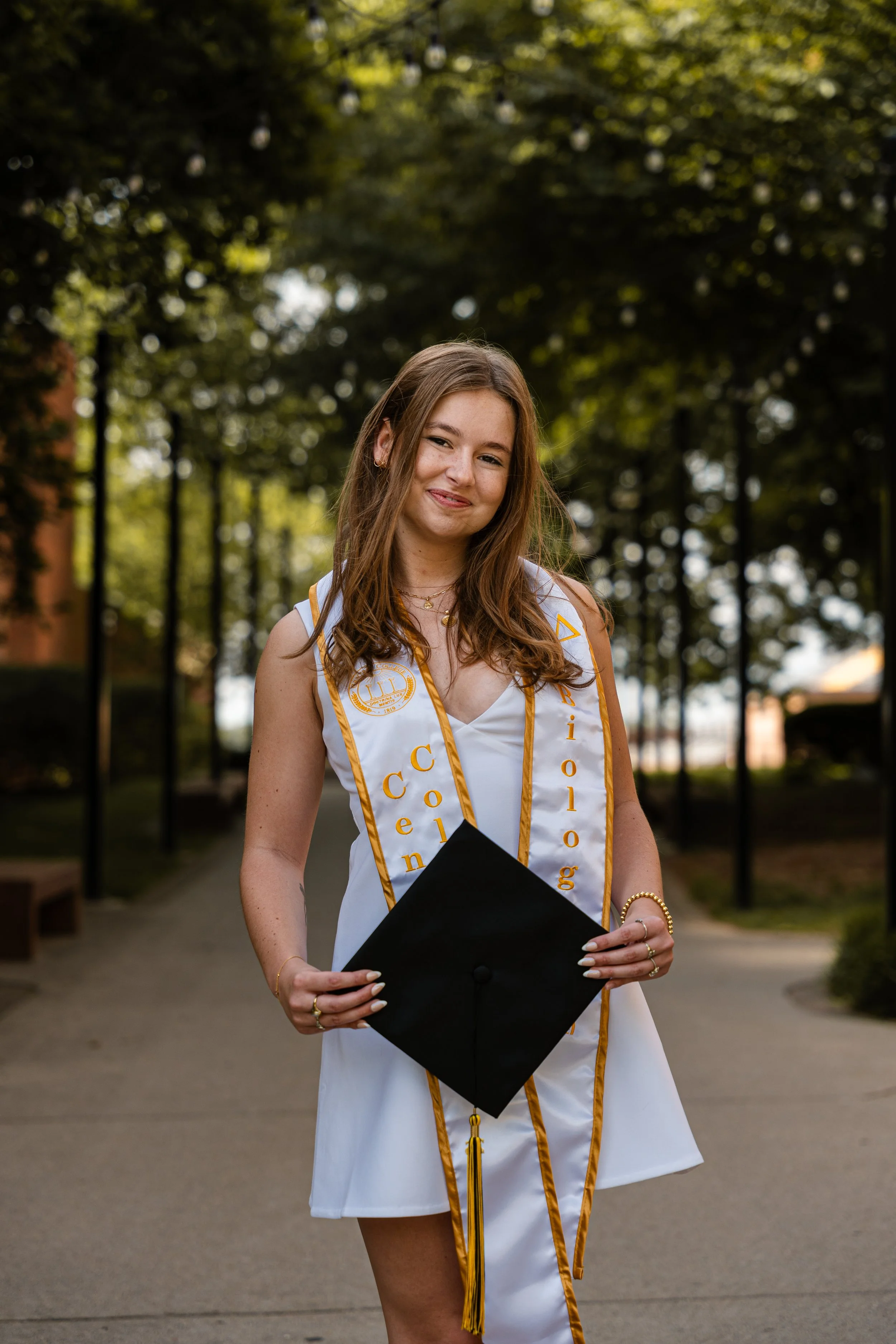 Young woman in a graduation cap and gown, smiling outdoors, holding her diploma, with a graduation stole that reads 'Congratulations' and leopard print earrings.