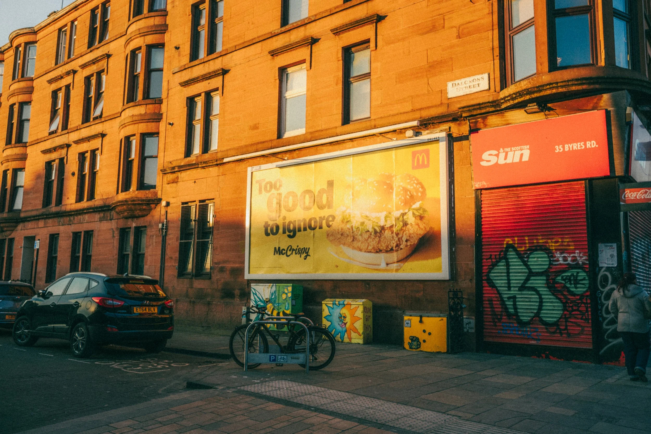 Street scene showing a multi-story brick building with large windows, advertisements including a McDonald's billboard, parked cars, bicycles, and graffiti on a red shutter.