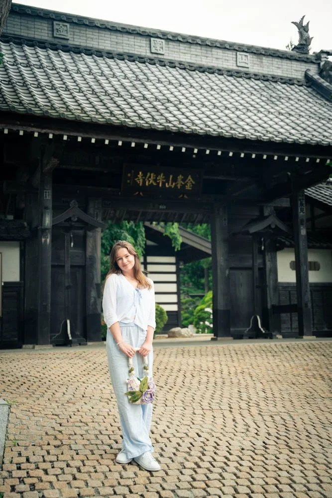 A woman standing in front of a traditional Japanese temple gate, holding a small floral handbag, with greenery visible behind the gate.