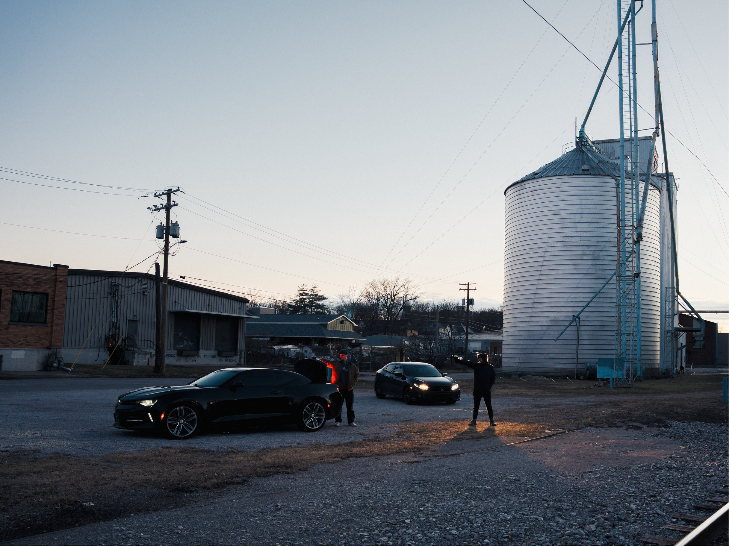 A parking lot during dusk with two black cars parked, a man and a woman standing near the cars, a large white grain silo with a ladder and metal structure on it, utility poles with wires, buildings, leafless trees, and a cloudy sky.