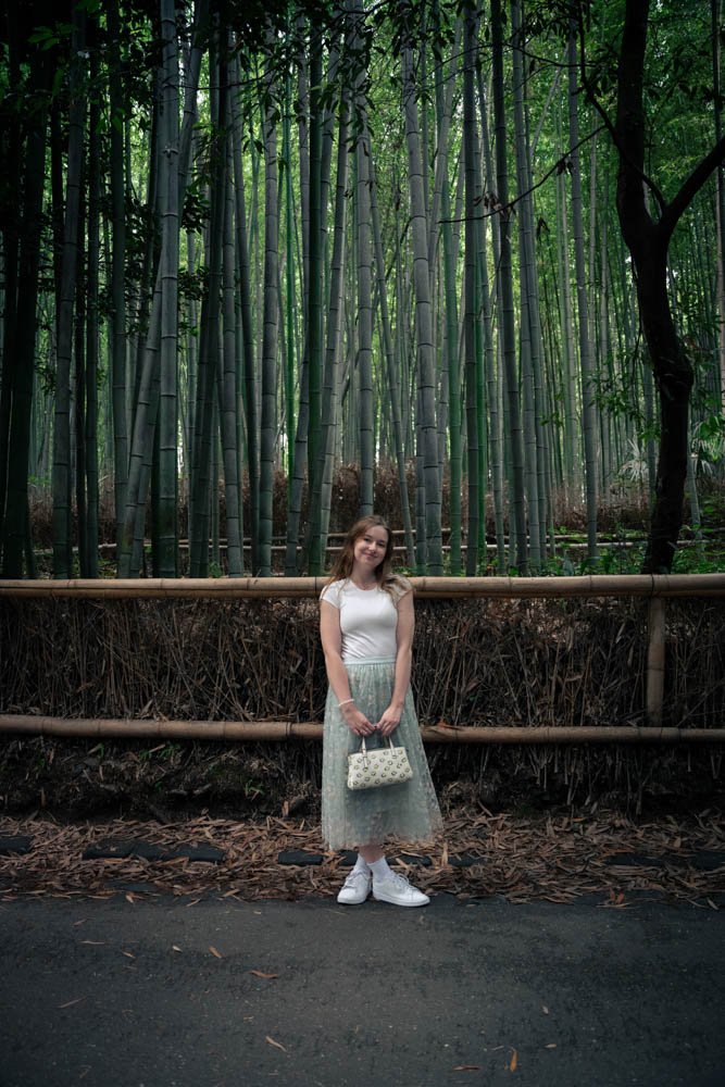A young woman standing in front of a bamboo forest, holding a small handbag, wearing a white top, a pastel skirt, and white sneakers.