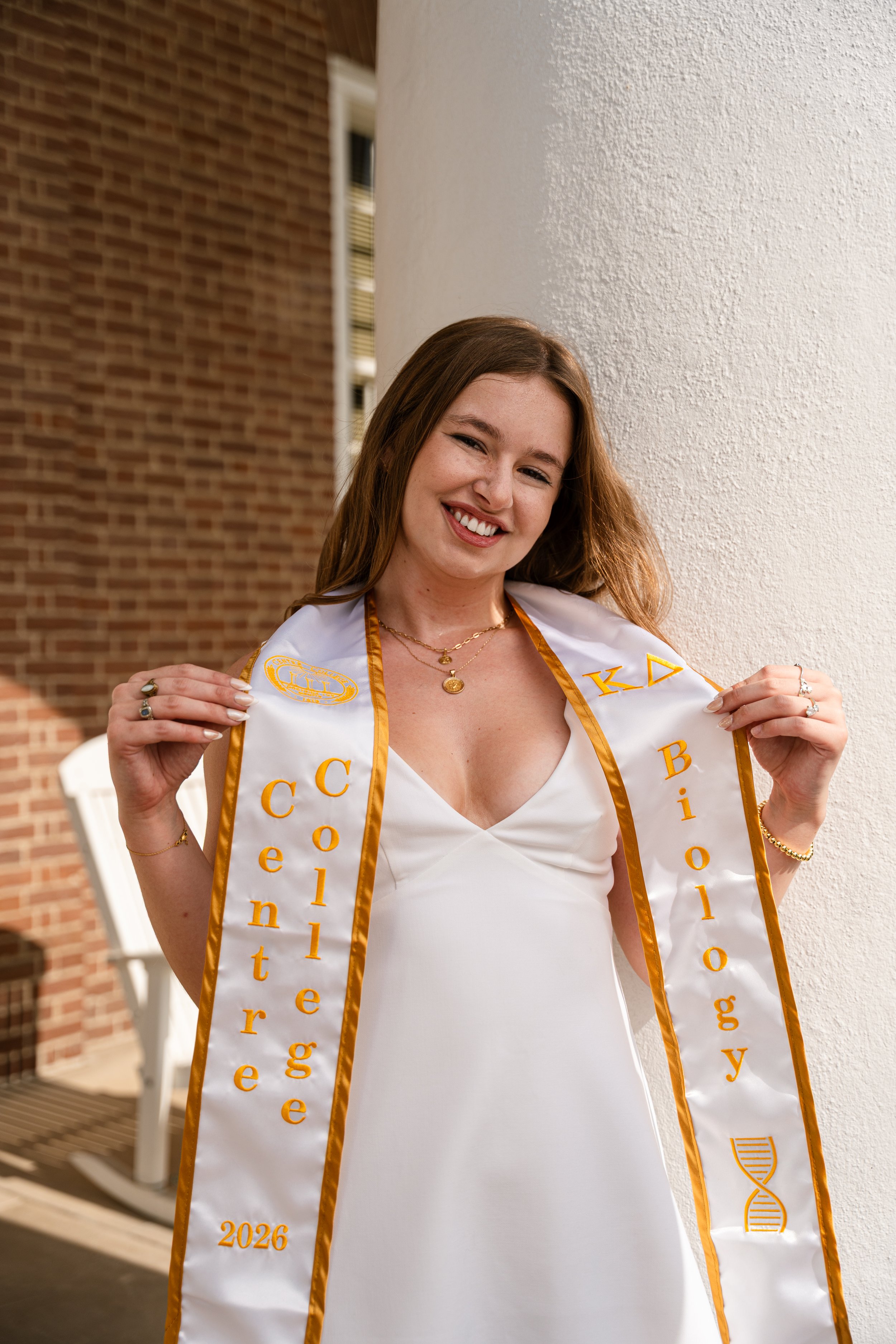 A young woman with brown hair smiling, wearing a white dress and a white sash with orange Greek letters, standing outside near a brick wall and a white column, holding the sash at her shoulders.