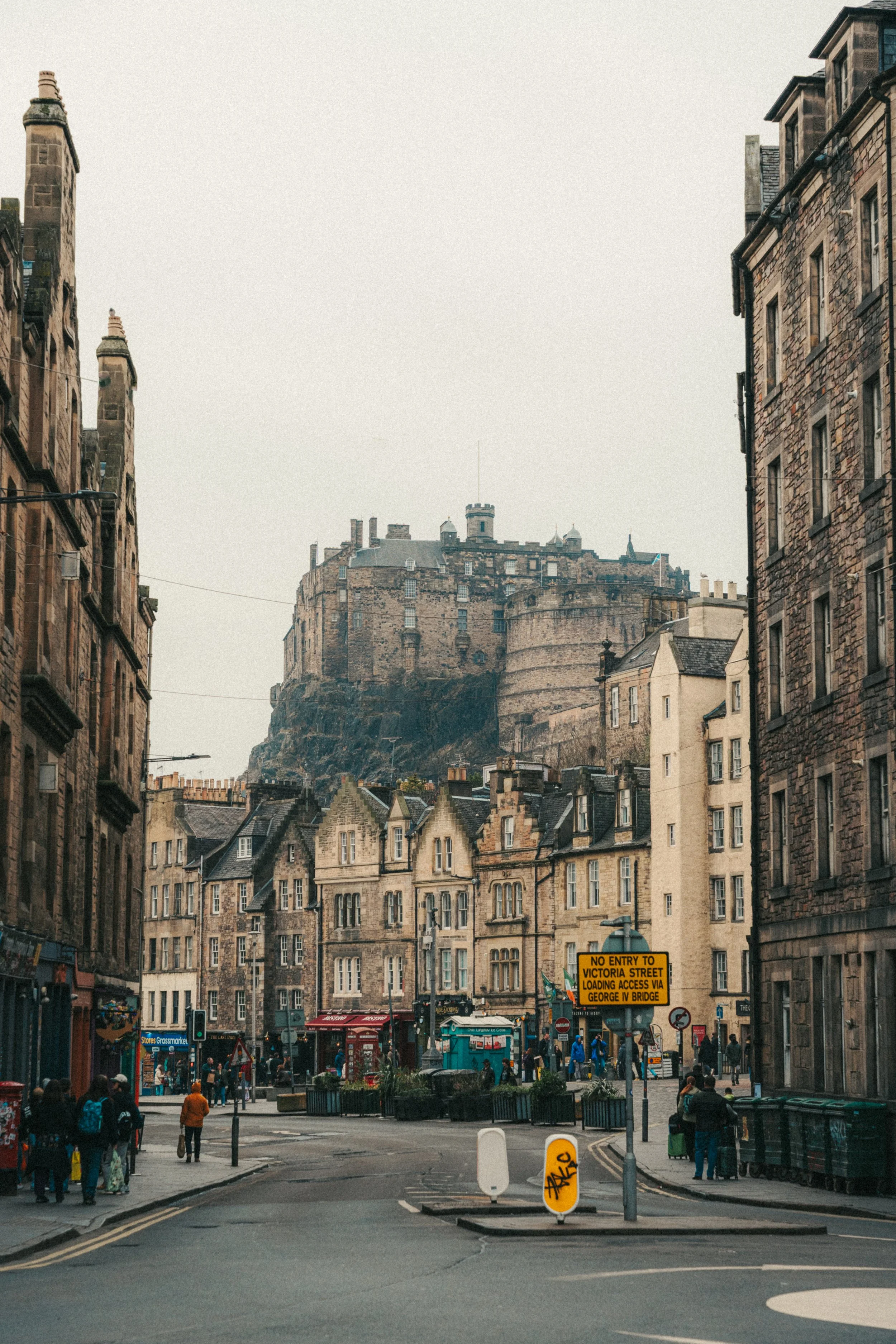 A busy urban street scene with historic stone buildings, a castle on a hill in the background, and pedestrians crossing at a city intersection. There are storefronts, street signs, and a mix of modern and historic architecture.