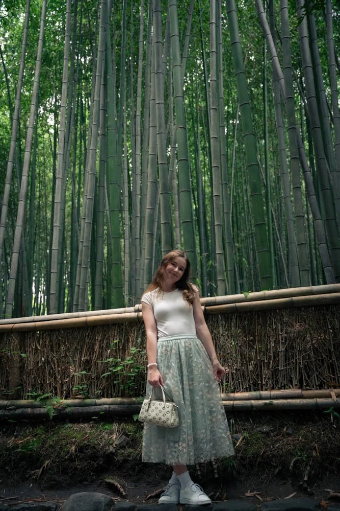 A woman standing in front of a green bamboo forest, holding a white purse and wearing a white top, patterned skirt, and white sneakers.