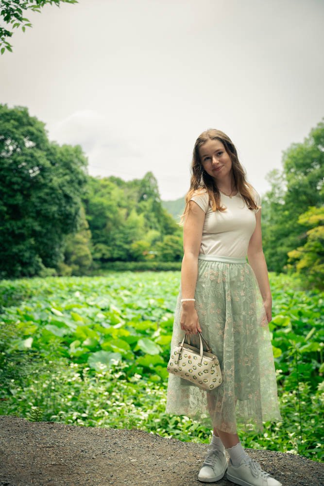 A young woman with long brown hair stands outdoors on a dirt path, holding a patterned bag in her right hand. She is dressed in a white top and a light green, lace skirt with a floral pattern. The background features a lush green landscape with trees