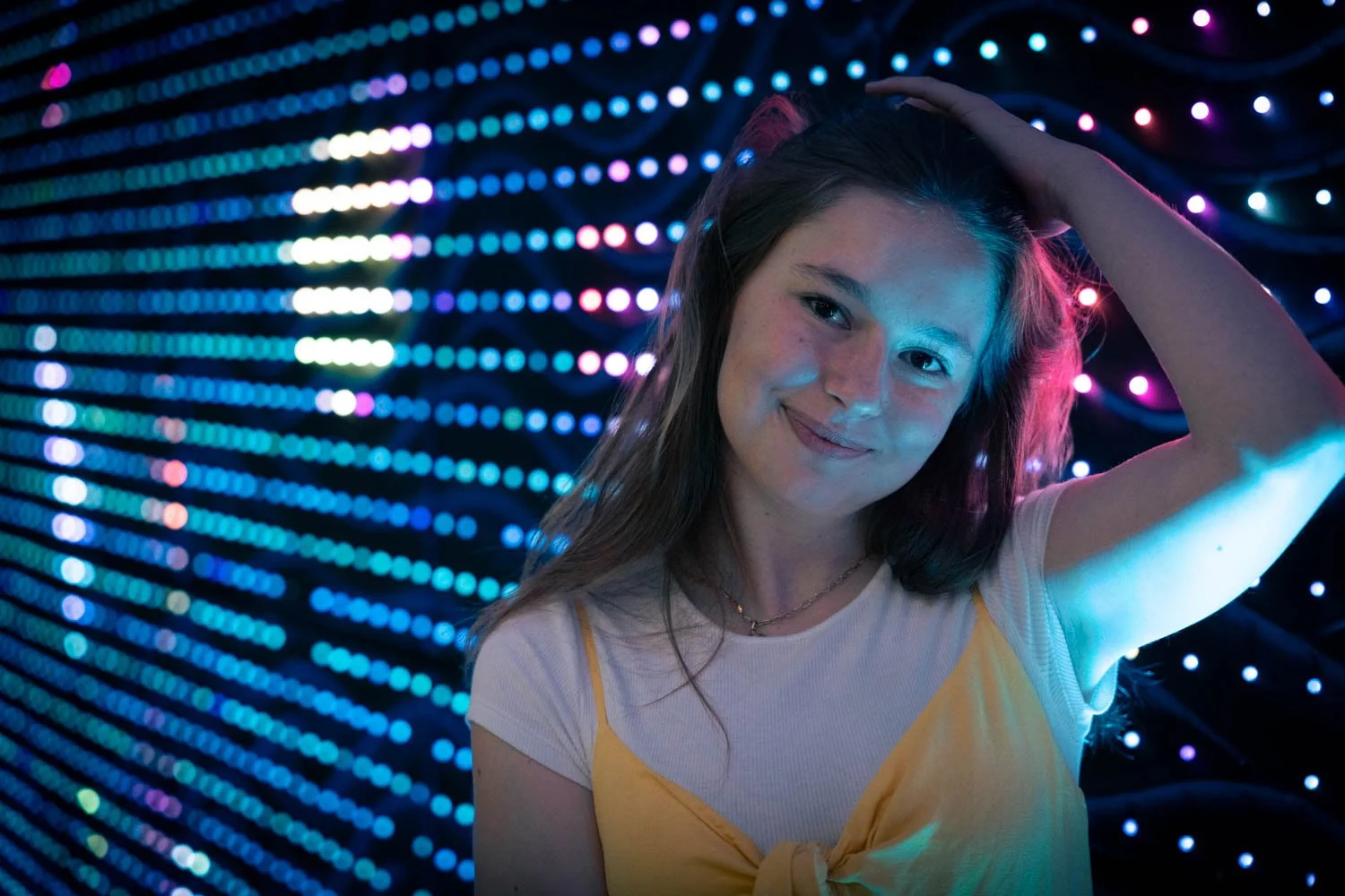 A young woman with long brown hair smiling and touching her head, with colorful, blurred lights in the background.