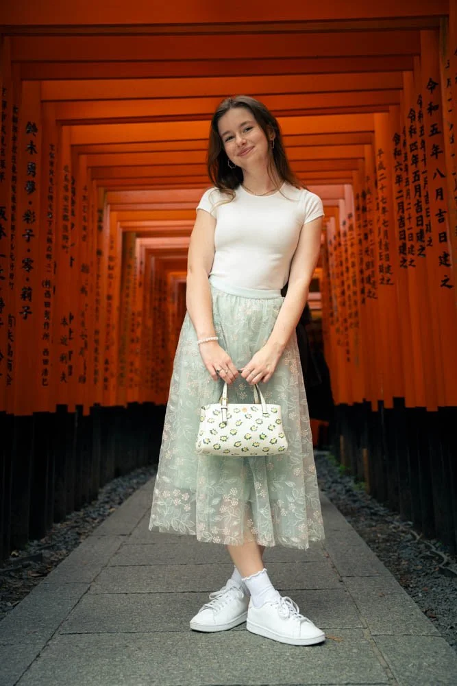 Young woman standing on a stone pathway, holding a white handbag with a lemon print, in front of a tunnel of orange torii gates with Japanese inscriptions.