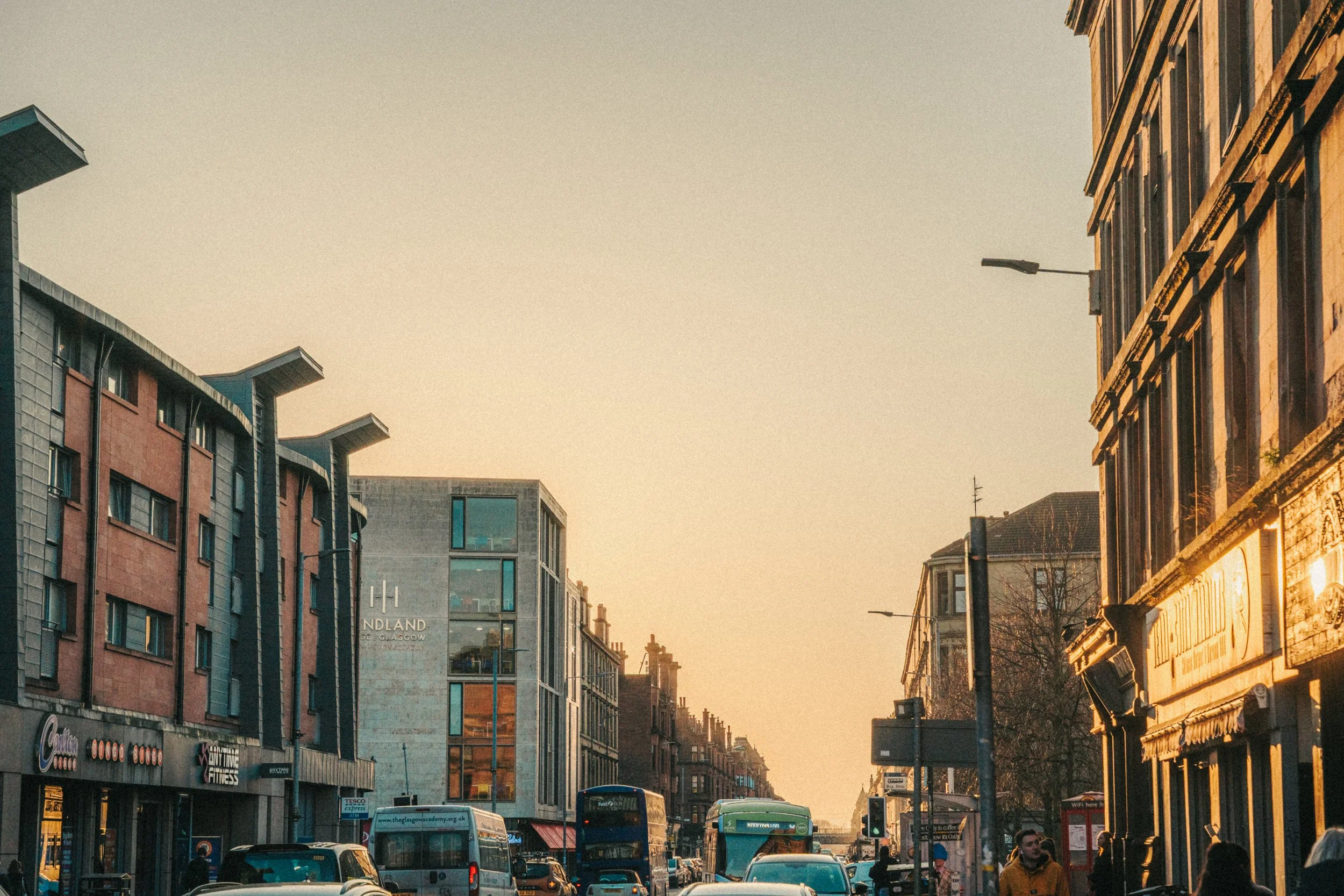 Street scene in an urban area with buildings, cars, buses, and pedestrians during sunset or golden hour.