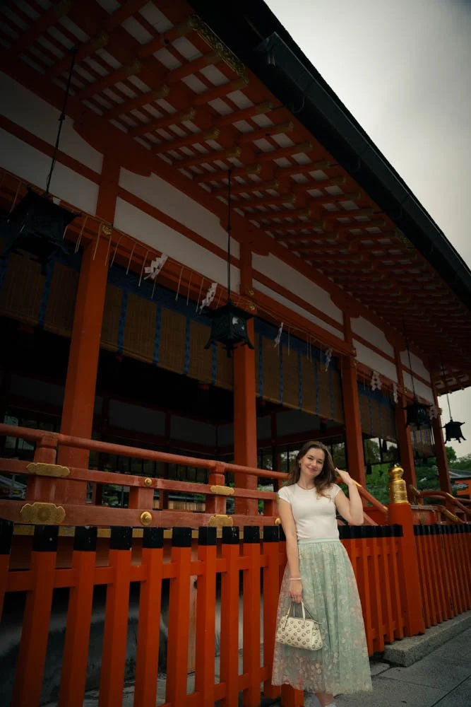 A woman in a white top and flowy skirt holding a patterned handbag, standing in front of a traditional Japanese temple with red wooden structure and decorative elements.