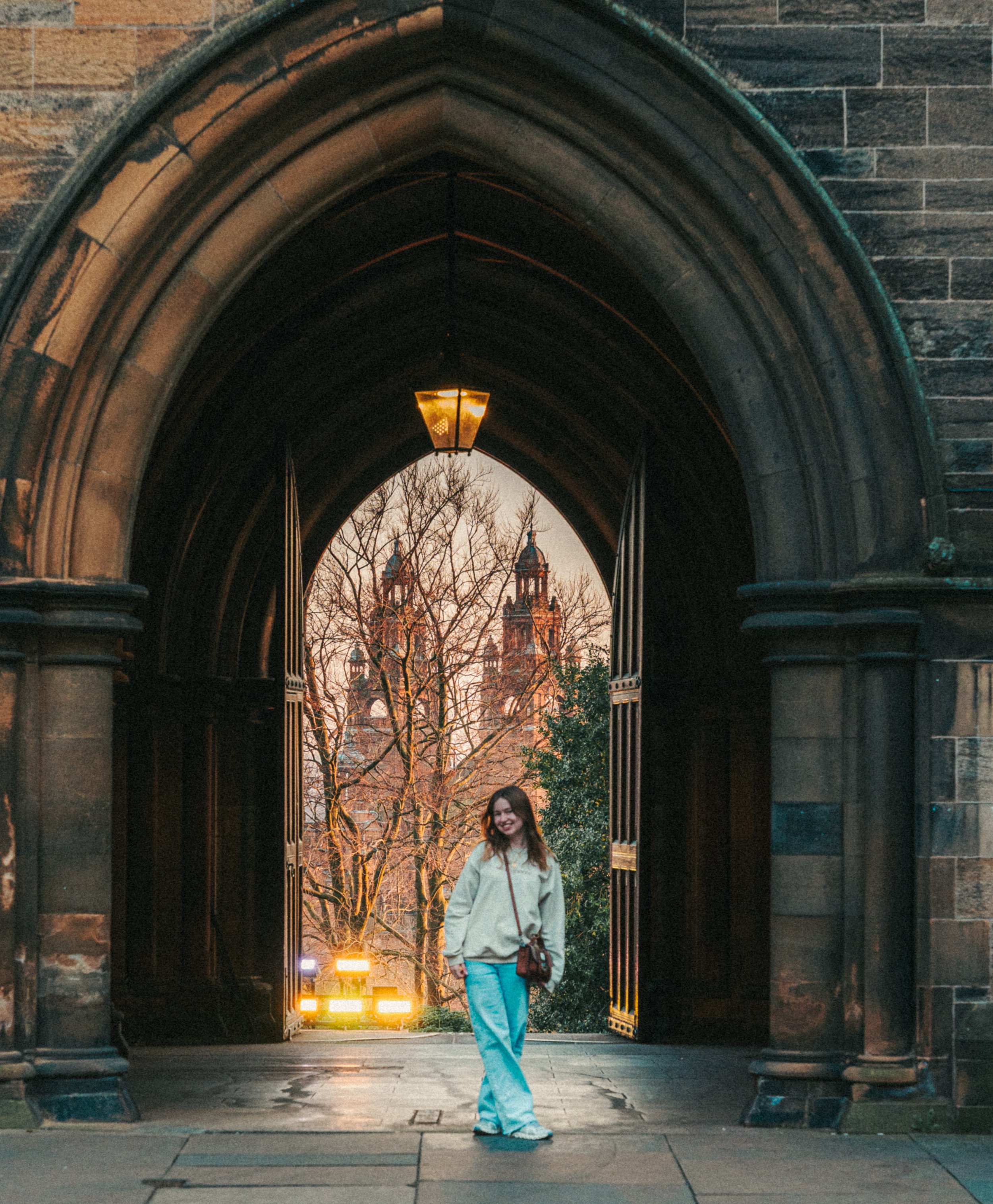 A young woman in light blue jeans, a gray zip-up hoodie, and white sneakers walking through a large stone archway with a lit lantern hanging from the ceiling. There are trees and a historical building with spires in the background, and streetlights i