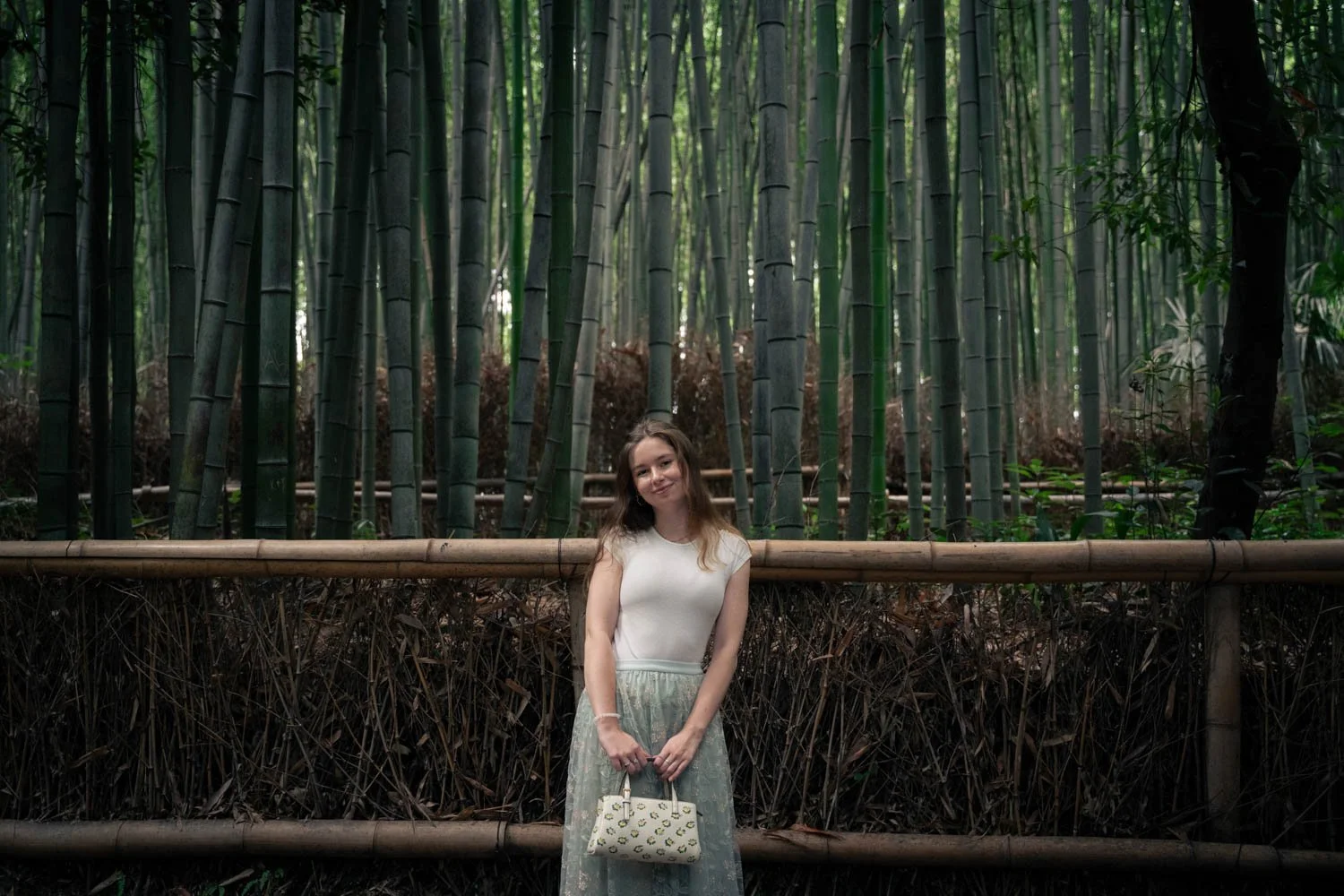A young woman standing in front of a bamboo forest, holding a patterned handbag, smiling softly.