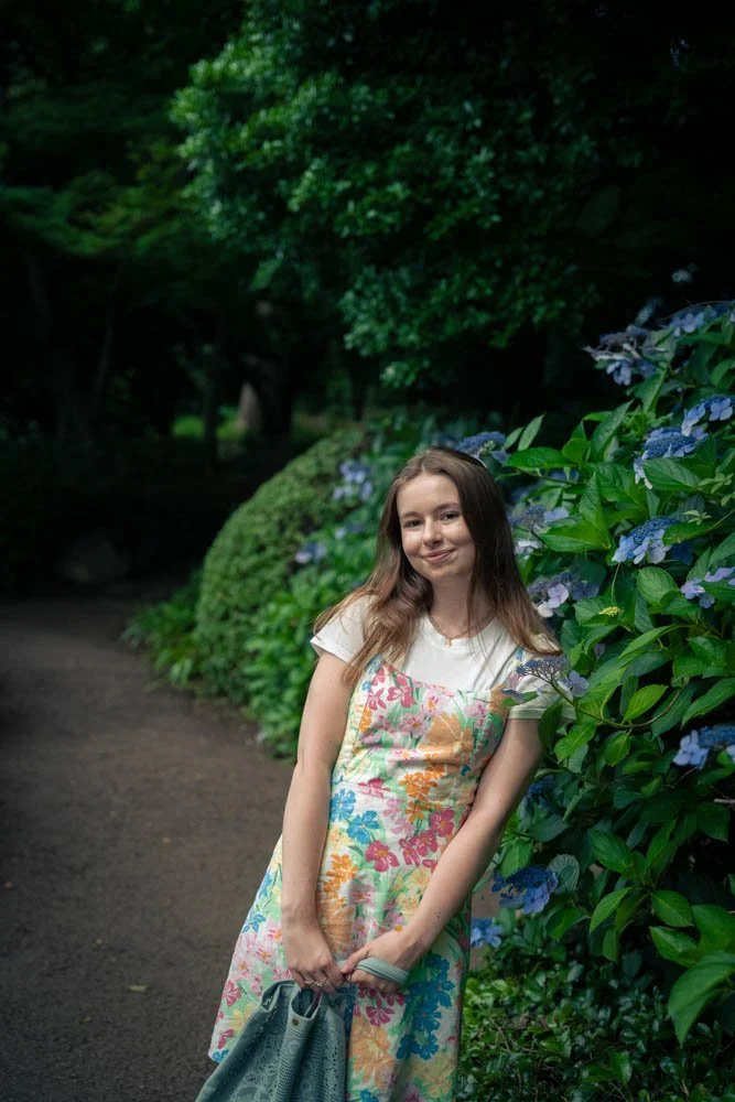 A young woman standing on a garden path next to a bush with purple flowers, surrounded by lush green trees and foliage.