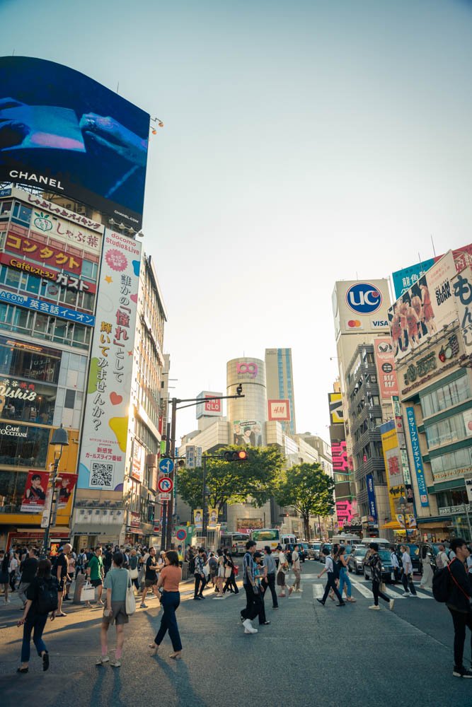 Busy crosswalk in a city with tall buildings, billboards, and advertisements, including Chanel and UNIQLO, in bright daylight.