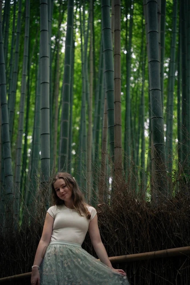 Young woman with brown hair, wearing a white top and floral skirt, sitting on a wooden railing in a dense bamboo forest.
