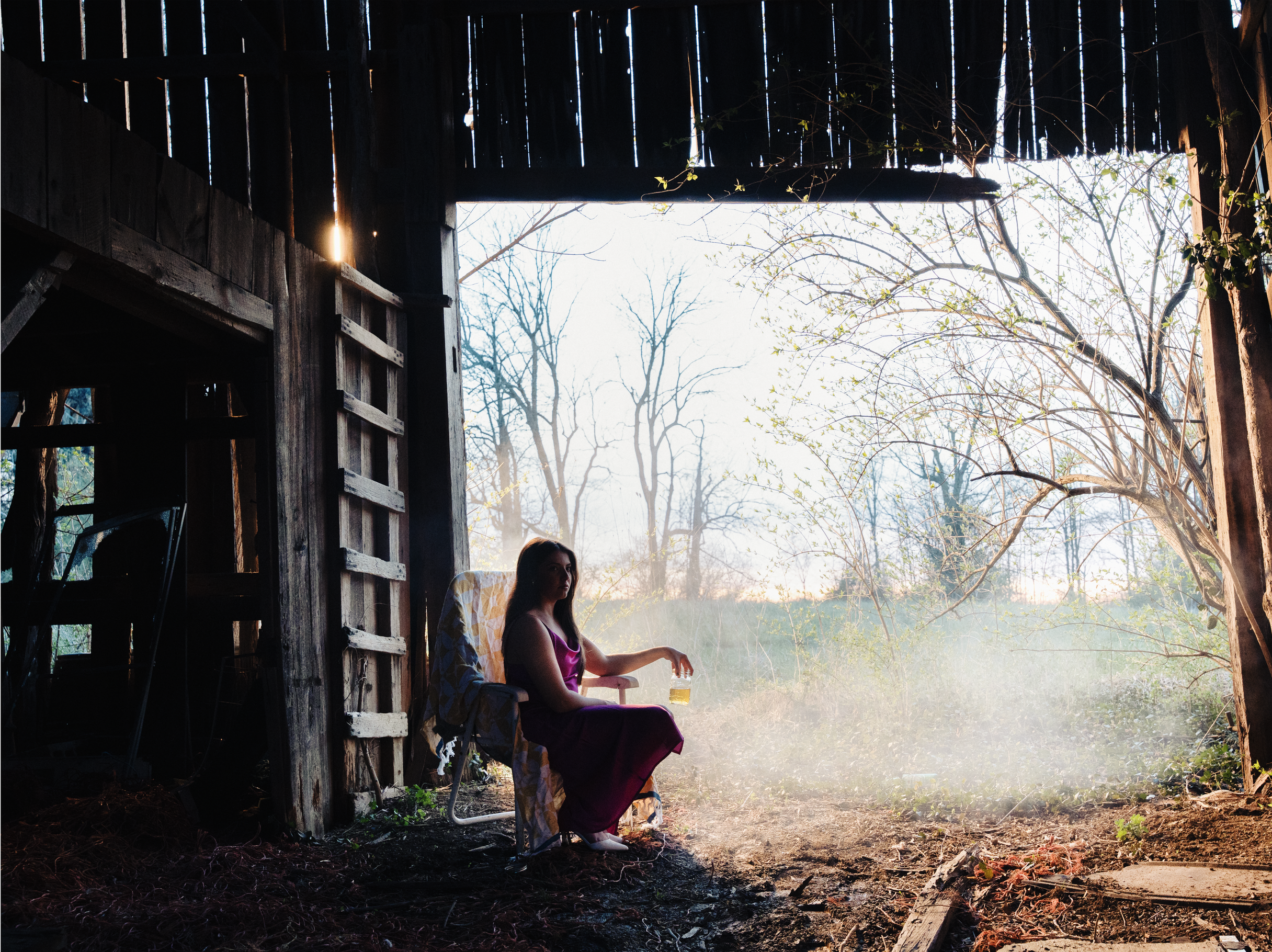 A woman sitting on a folding chair outside of a wooden barn, holding a bottle of beer, with sunlight streaming through the trees behind her.