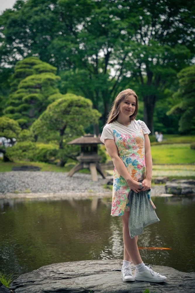 A young girl with long hair standing on a rock by a pond in a lush, green garden with trees and trimmed bushes, holding a jacket, wearing a colorful dress and white sneakers.