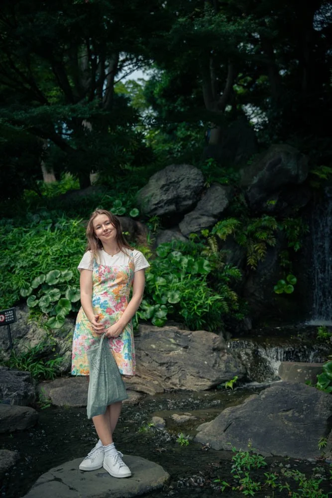A young woman standing on a rock in a lush garden with a small waterfall and large rocks behind her, surrounded by green plants and trees.