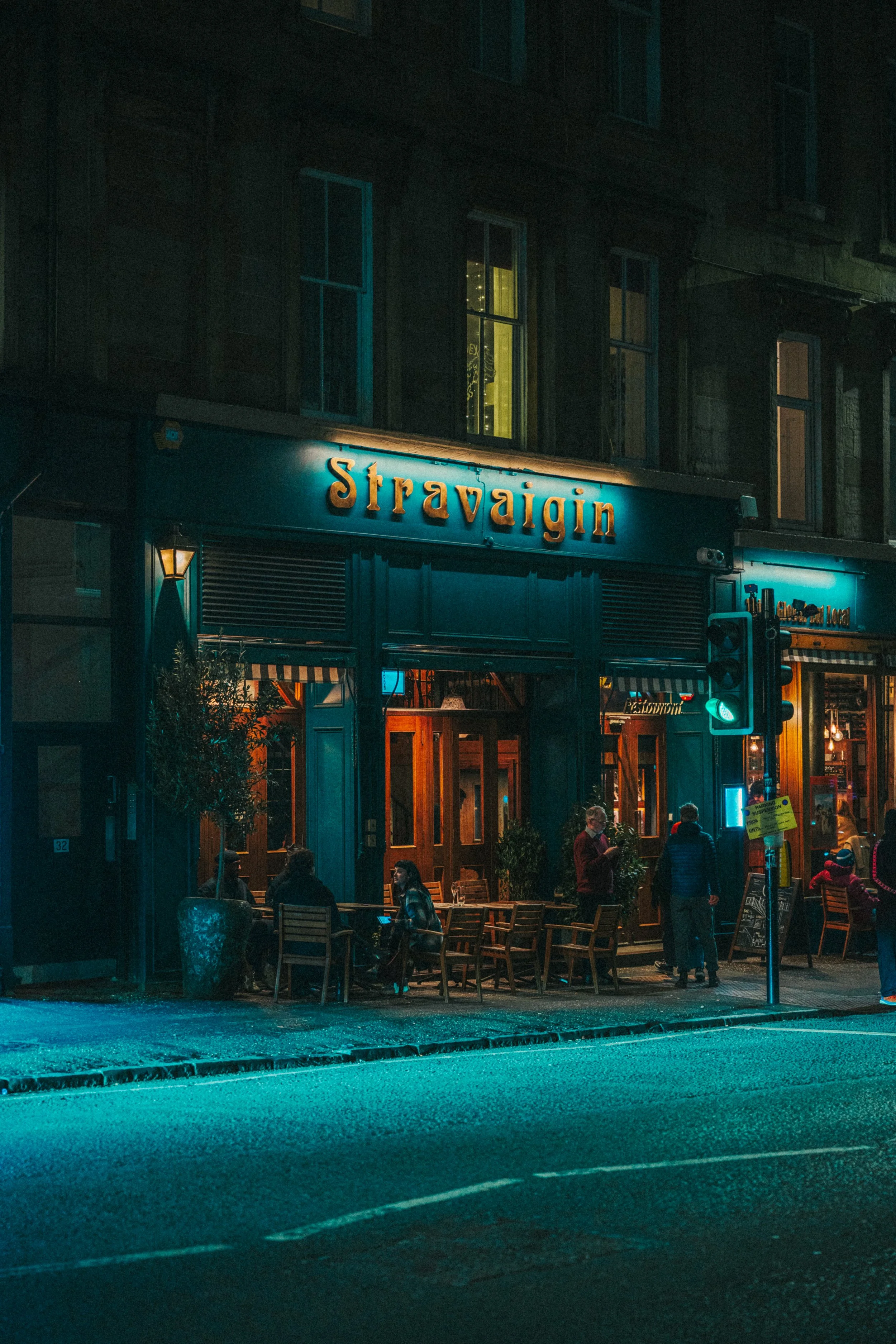 Nighttime scene outside a restaurant named 'Stravaigin' with people sitting at outdoor tables and others standing nearby, illuminated by street and restaurant lighting.