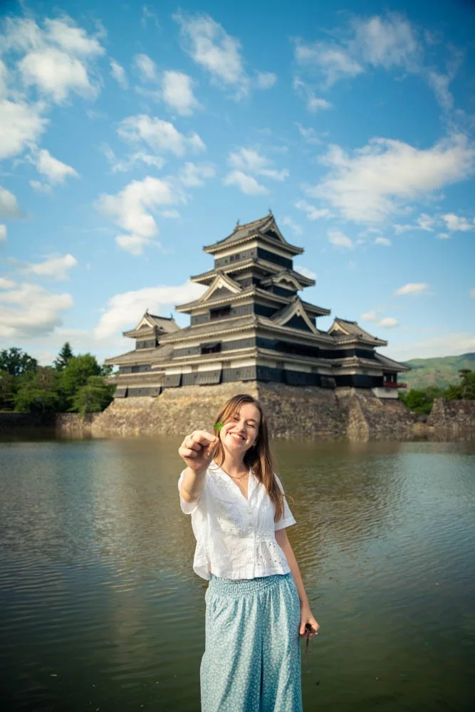 A smiling young woman holding a green leaf, standing in front of a traditional Japanese castle surrounded by water, under a partly cloudy sky.