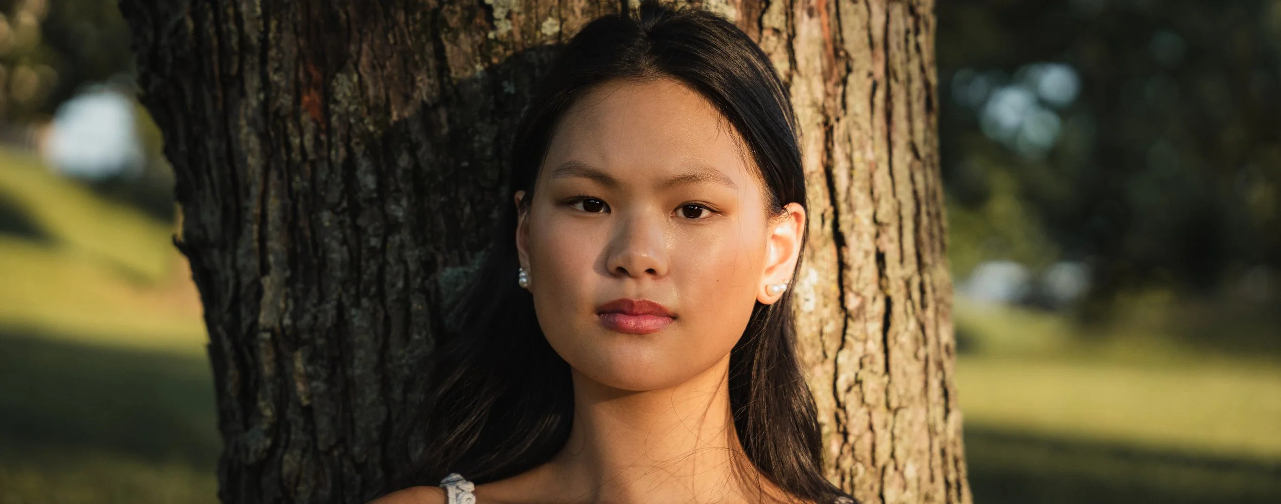 A young woman with long dark hair leaning against a tree trunk outdoors during late afternoon or early evening, with sunlight illuminating her face. She is wearing pearl earrings and a white top.