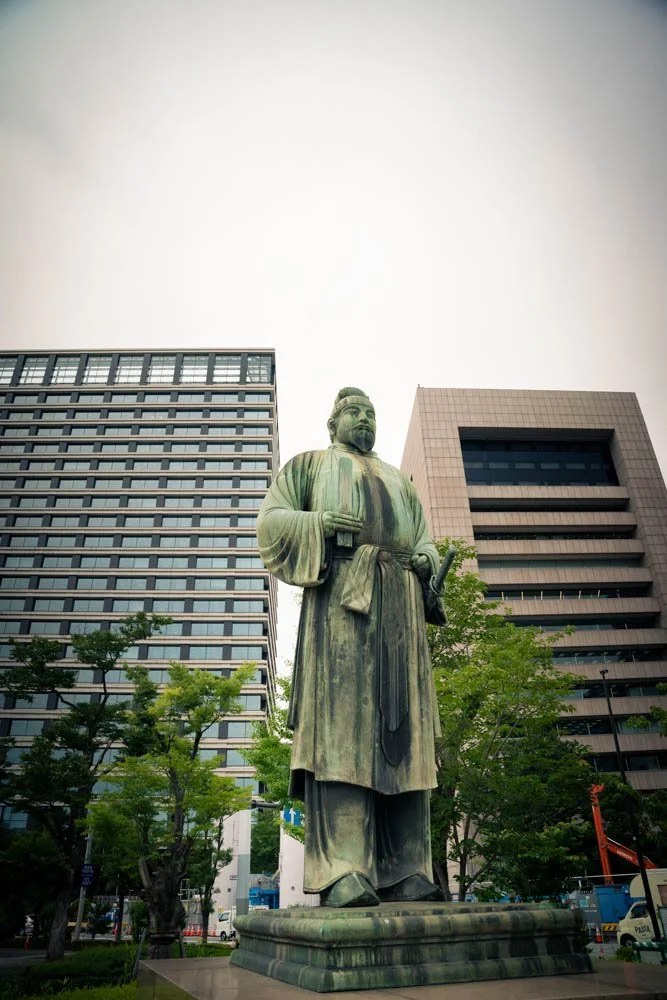 A large statue of a historical figure dressed in traditional clothing, standing outdoors in an urban setting with modern buildings and trees in the background.