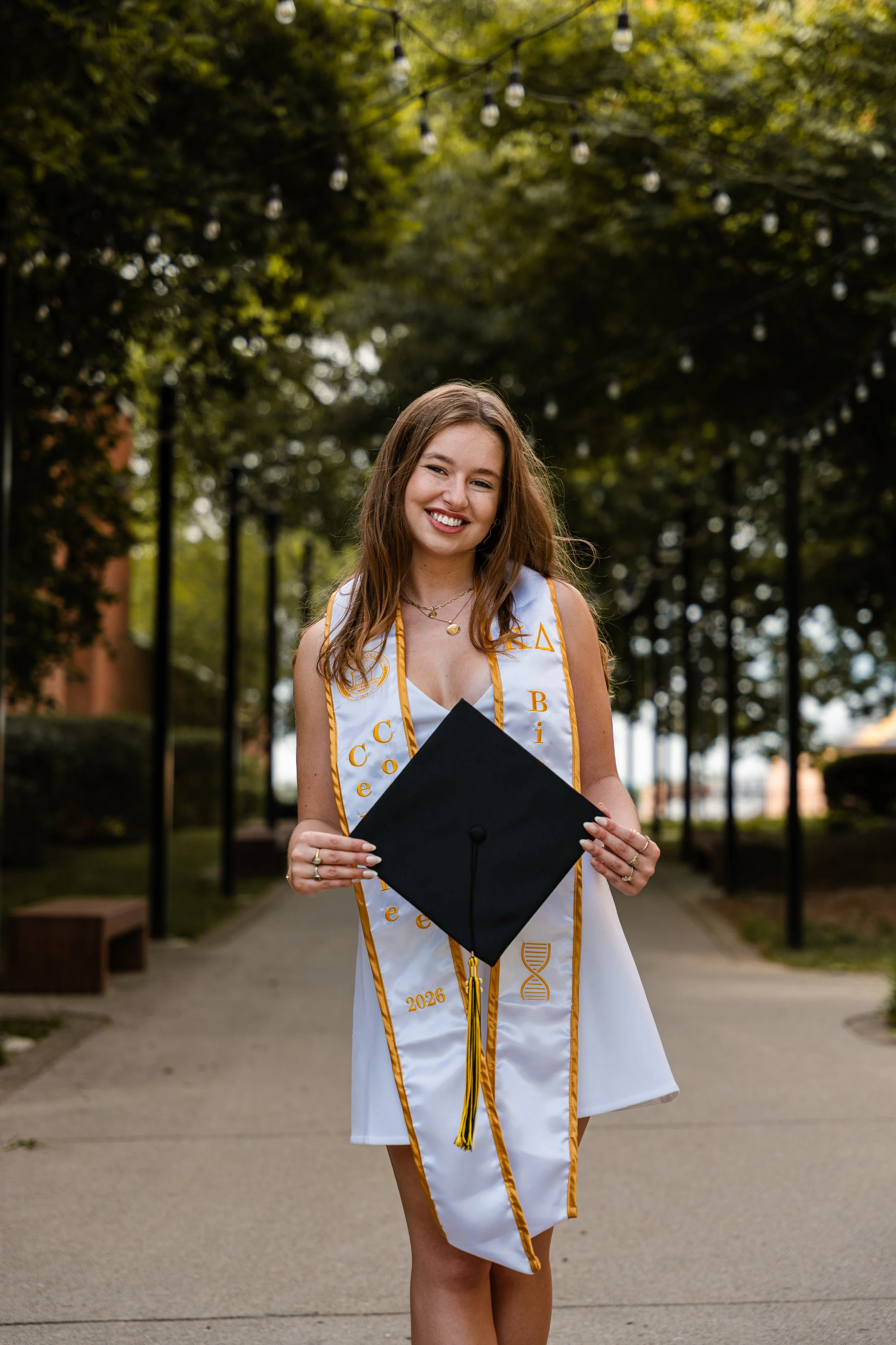 A young woman in a white graduation dress holding a black diploma cover and wearing a white honor stole with gold trim and Greek letters standing outdoors under string lights, smiling.