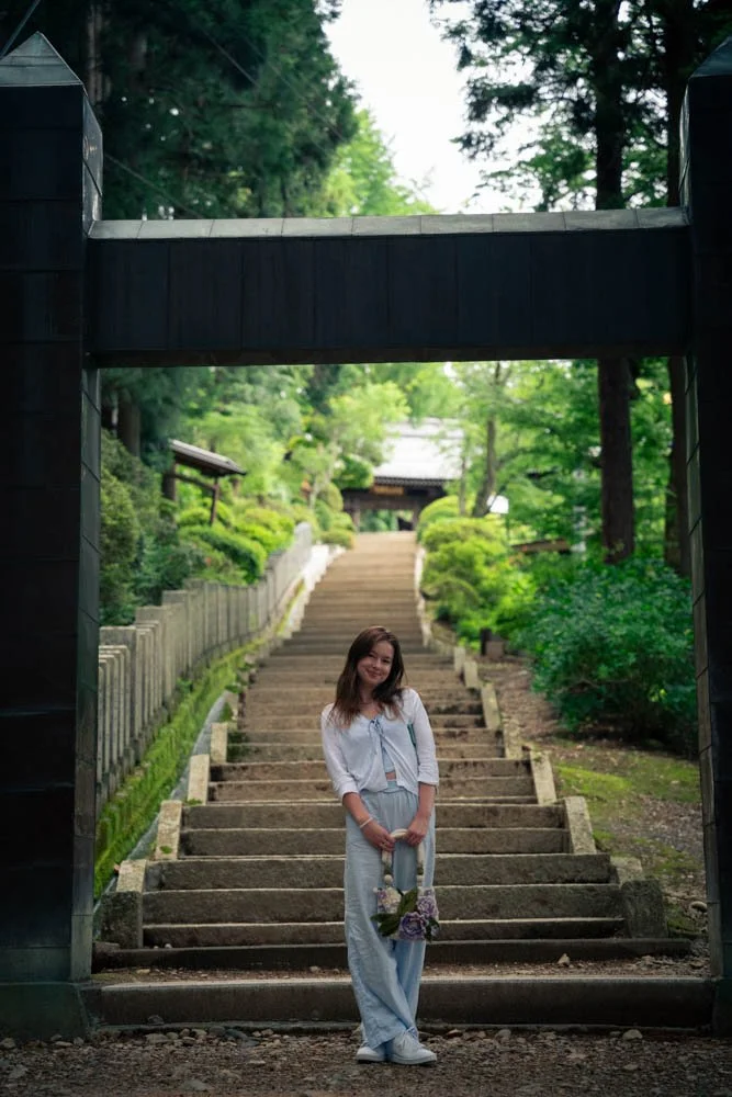 A young woman stands at the bottom of stone steps in a lush, green garden, holding a bouquet of purple flowers and smiling.