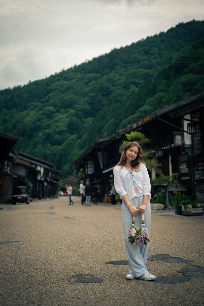 Young woman standing on a street in a mountain village, holding a rope with flowers, surrounded by traditional buildings and green hills.