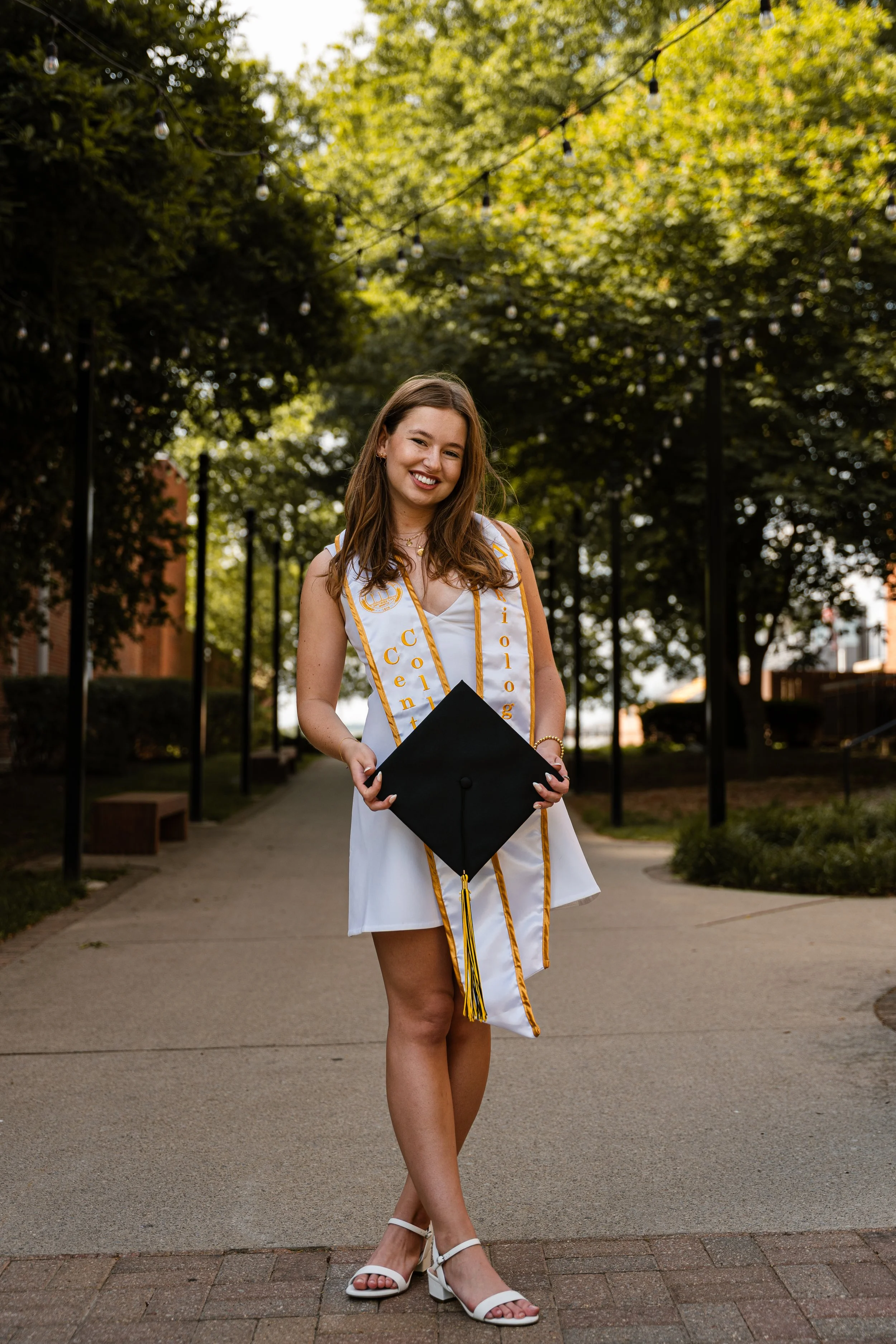 A young woman in a white dress and heels celebrating graduation, holding a black cap in one hand, with a tassel and wearing a white and gold stole, standing on a paved pathway in a park with green trees and hanging string lights in the background.