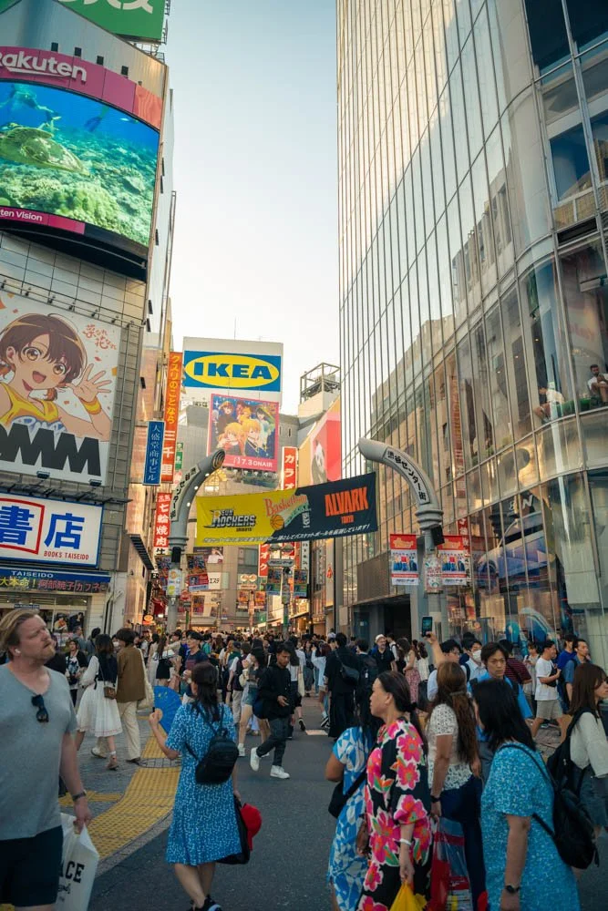 A busy city street scene in Japan with many pedestrians, tall buildings, and colorful billboards, including IKEA and anime advertisements.