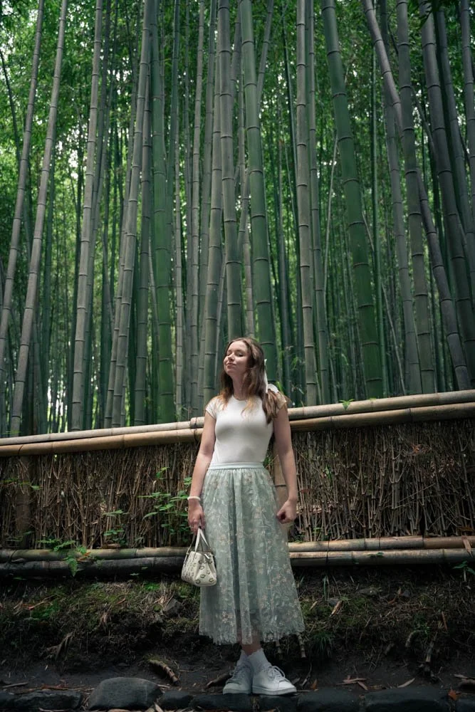 A woman in white top and lace skirt standing in front of a bamboo forest with tall green bamboo stalks.
