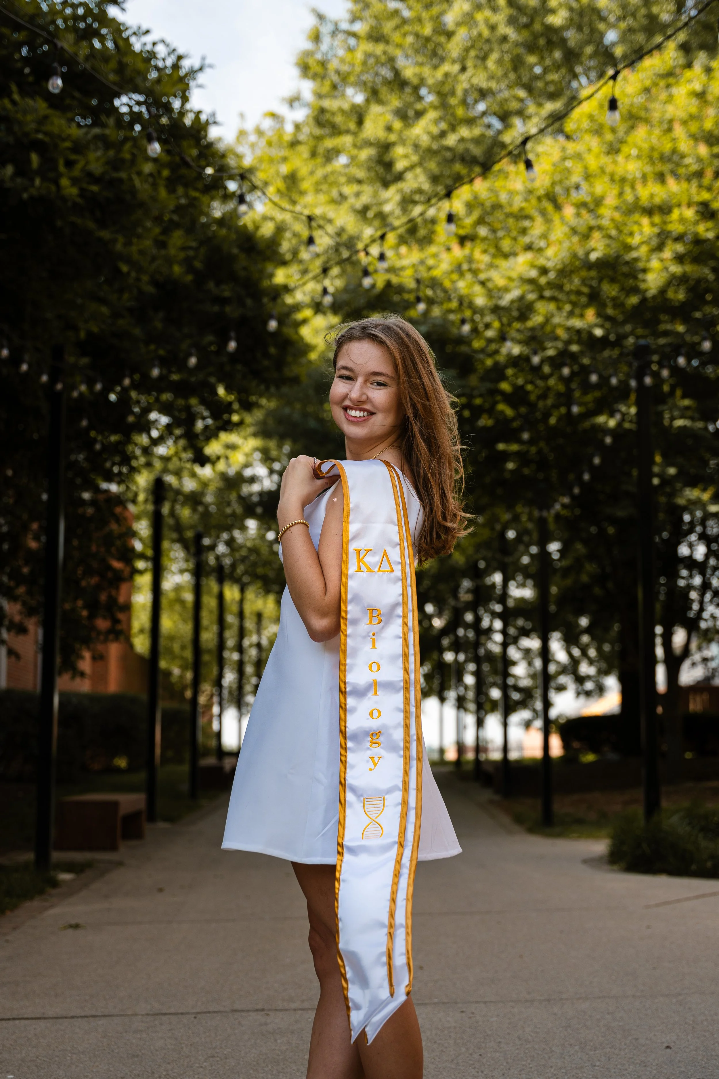 A young woman in a graduation gown smiling outdoors, holding her cap over her shoulder, with trees and string lights in the background.