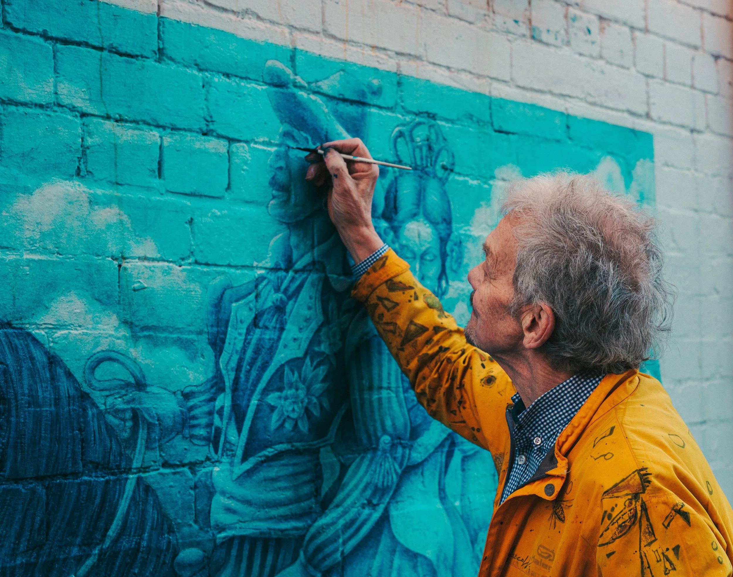 An elderly woman painting a mural on a brick wall, using blue paint, wearing a yellow jacket with black signatures, and a checkered shirt underneath.