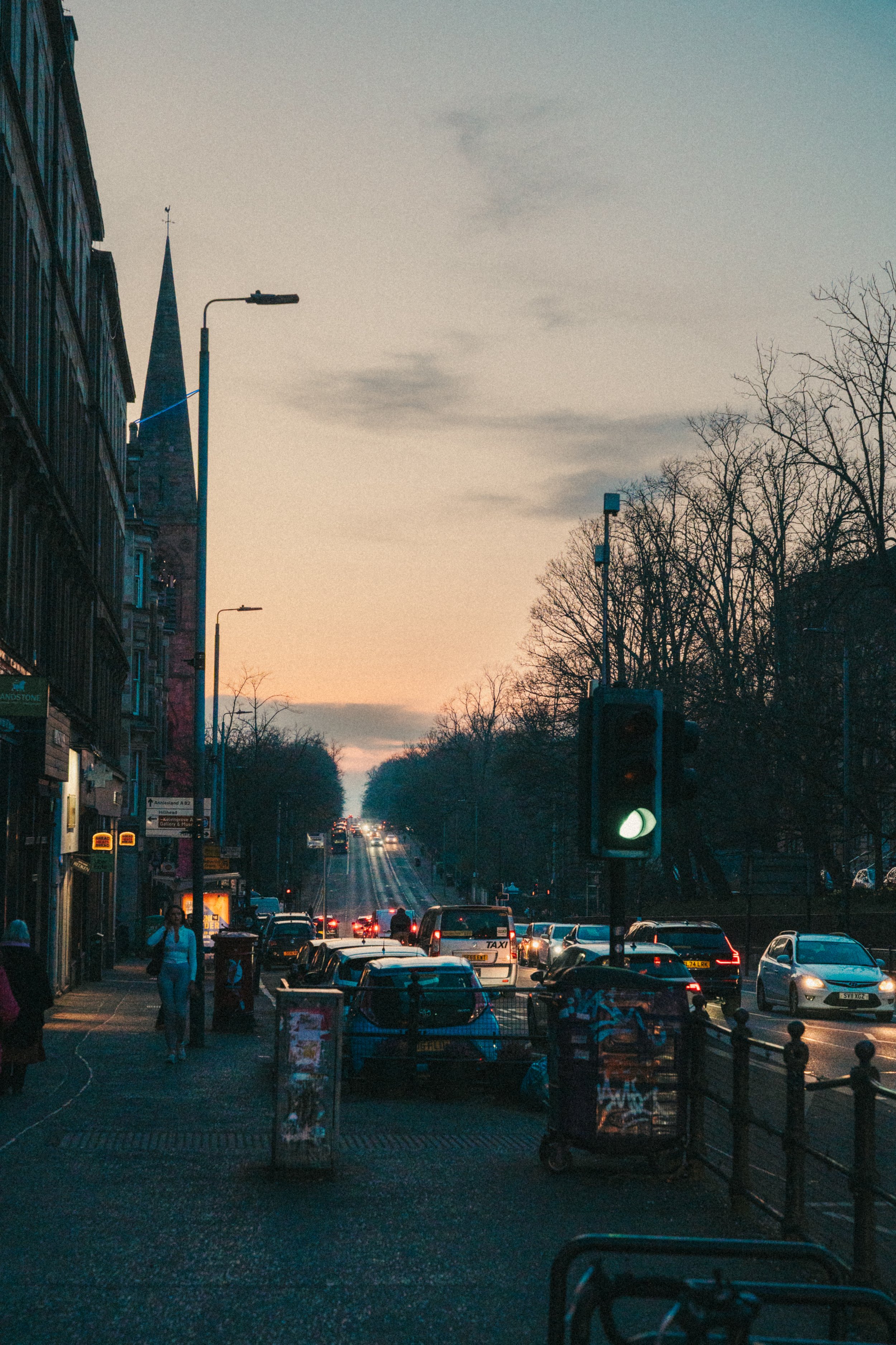 City street at dusk with cars and pedestrians, leafless trees, streetlights, and buildings, including a church steeple in the background.