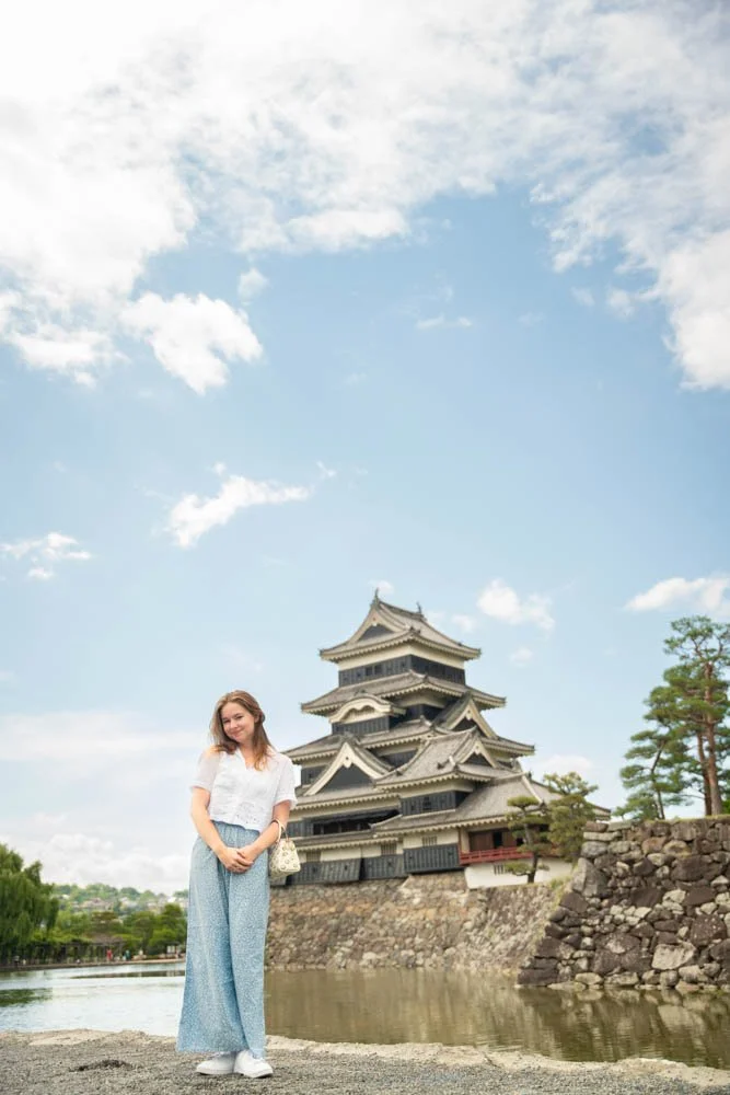 A young woman stands by a moat in front of a traditional Japanese castle under a partly cloudy sky.