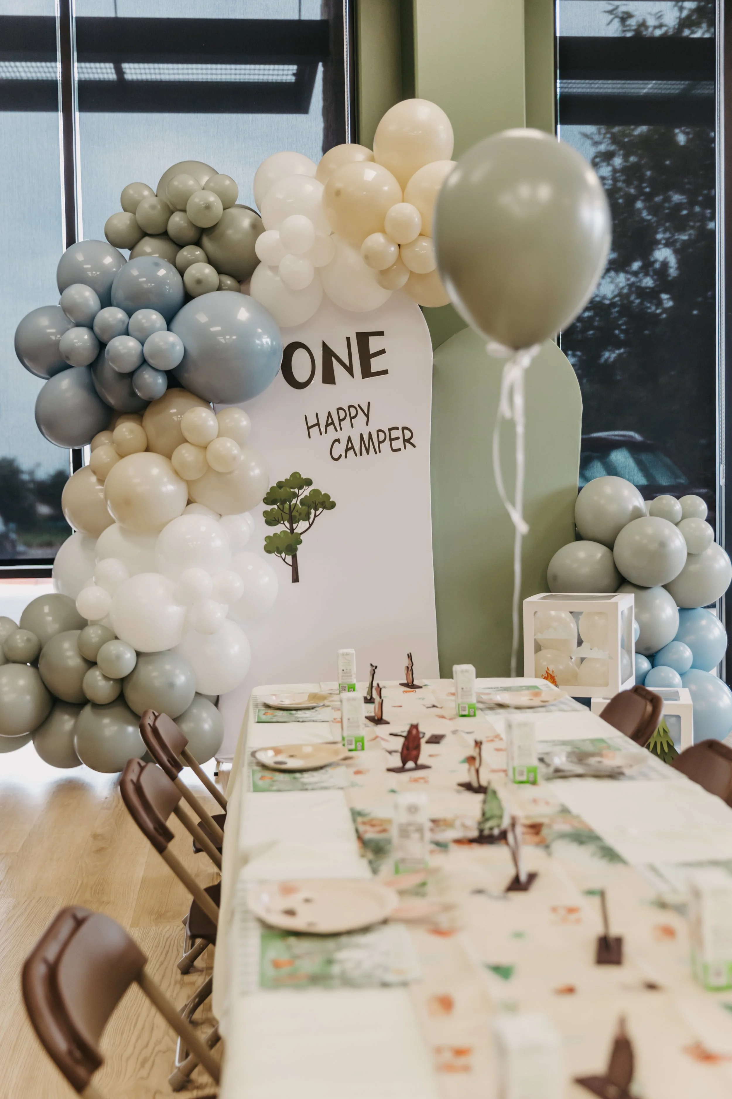 Indoor celebration setup with balloon decorations and a table with place settings, including a banner that reads 'ONE HAPPY CAMPER' and woodland-themed table decorations.