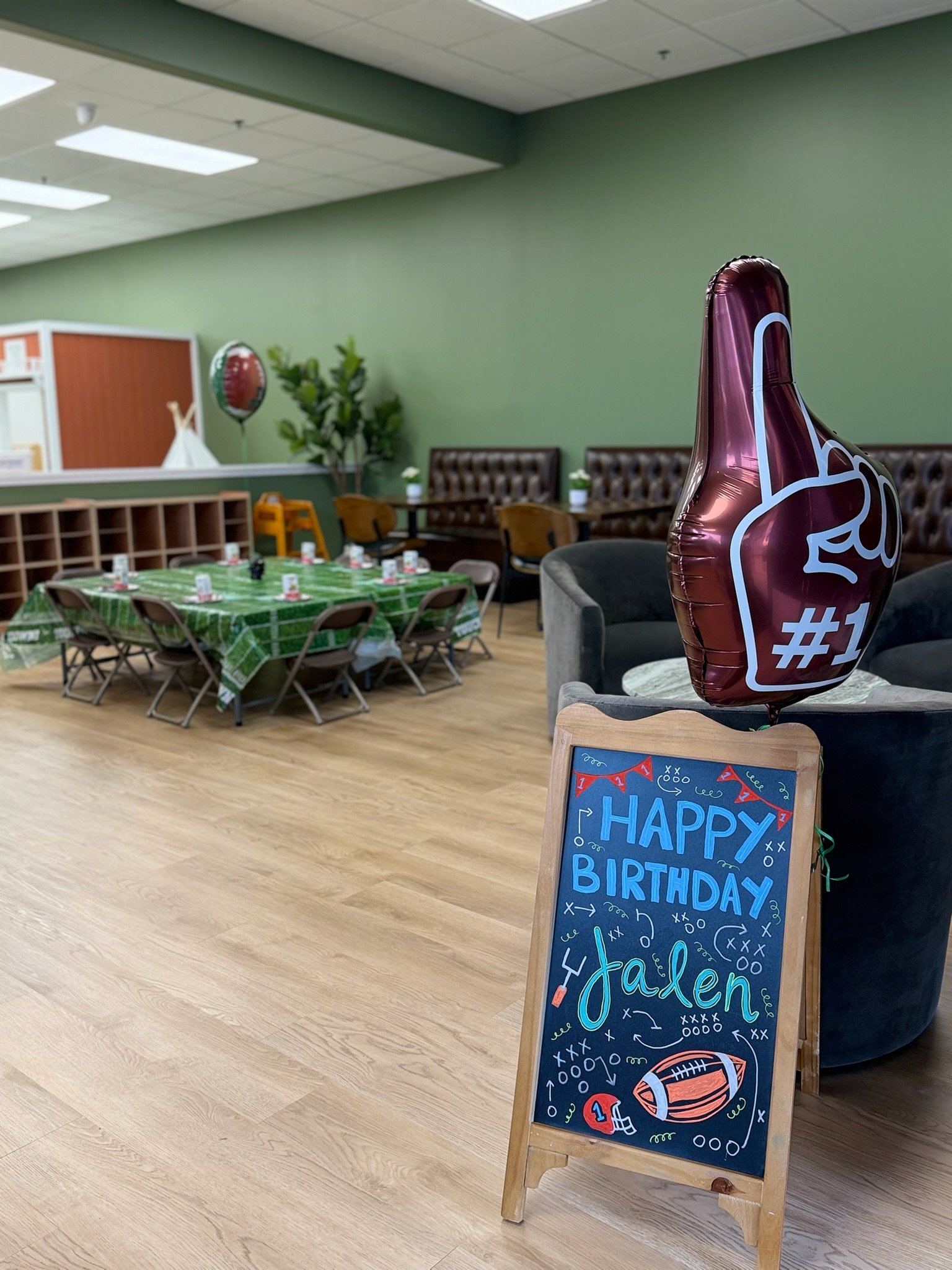 A decorated birthday party setup with a chalkboard sign that says 'Happy Birthday Ethan' and a foil balloon shaped like a thumbs-up with the #1 hand sign. The room has a green wall, brown chairs, and tables with green tablecloths. There are some small potted plants and a balloon in the background.