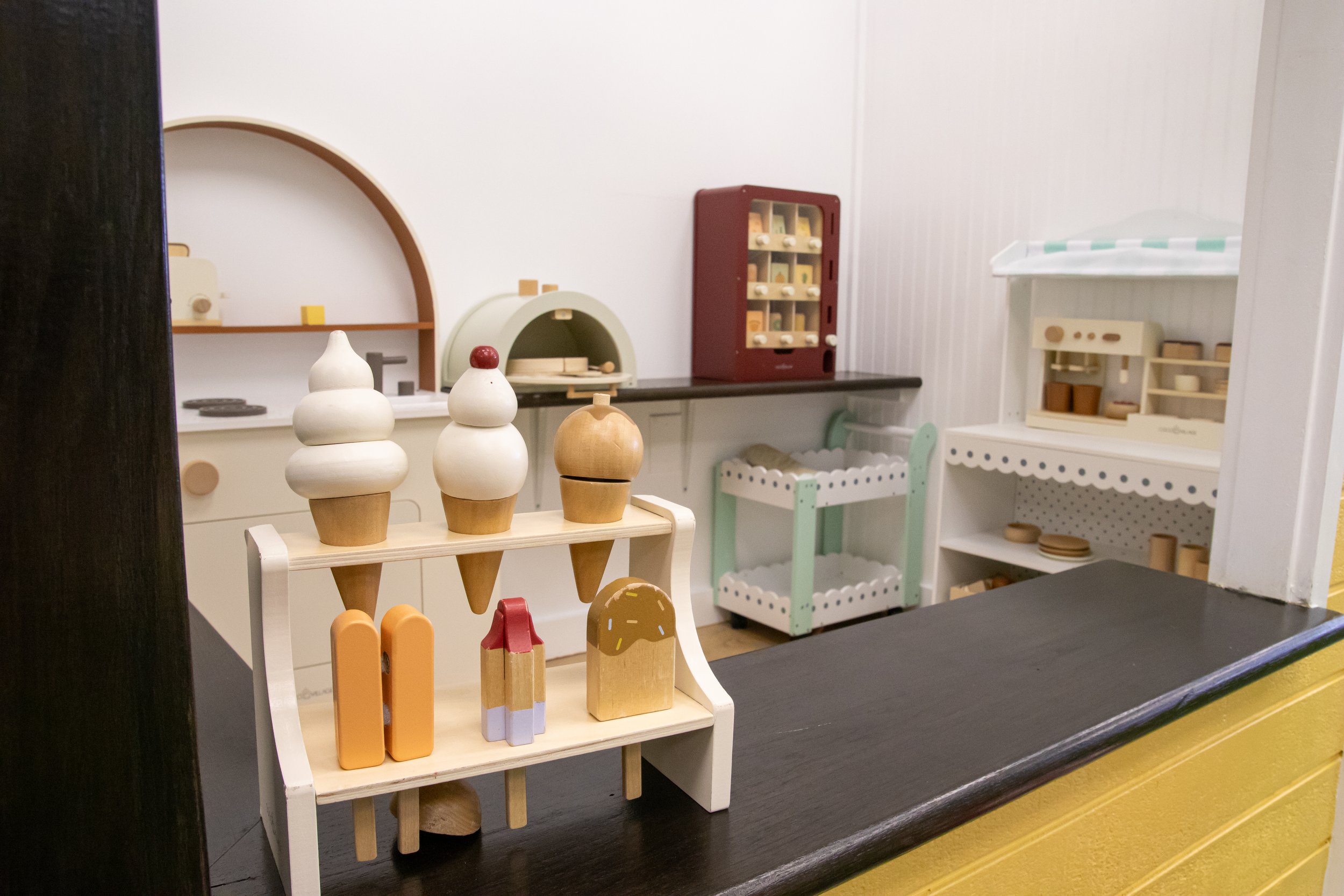 Miniature wooden toys on display in a toy store, including vases, popsicles, and various food items, with shelves holding more toys in the background.