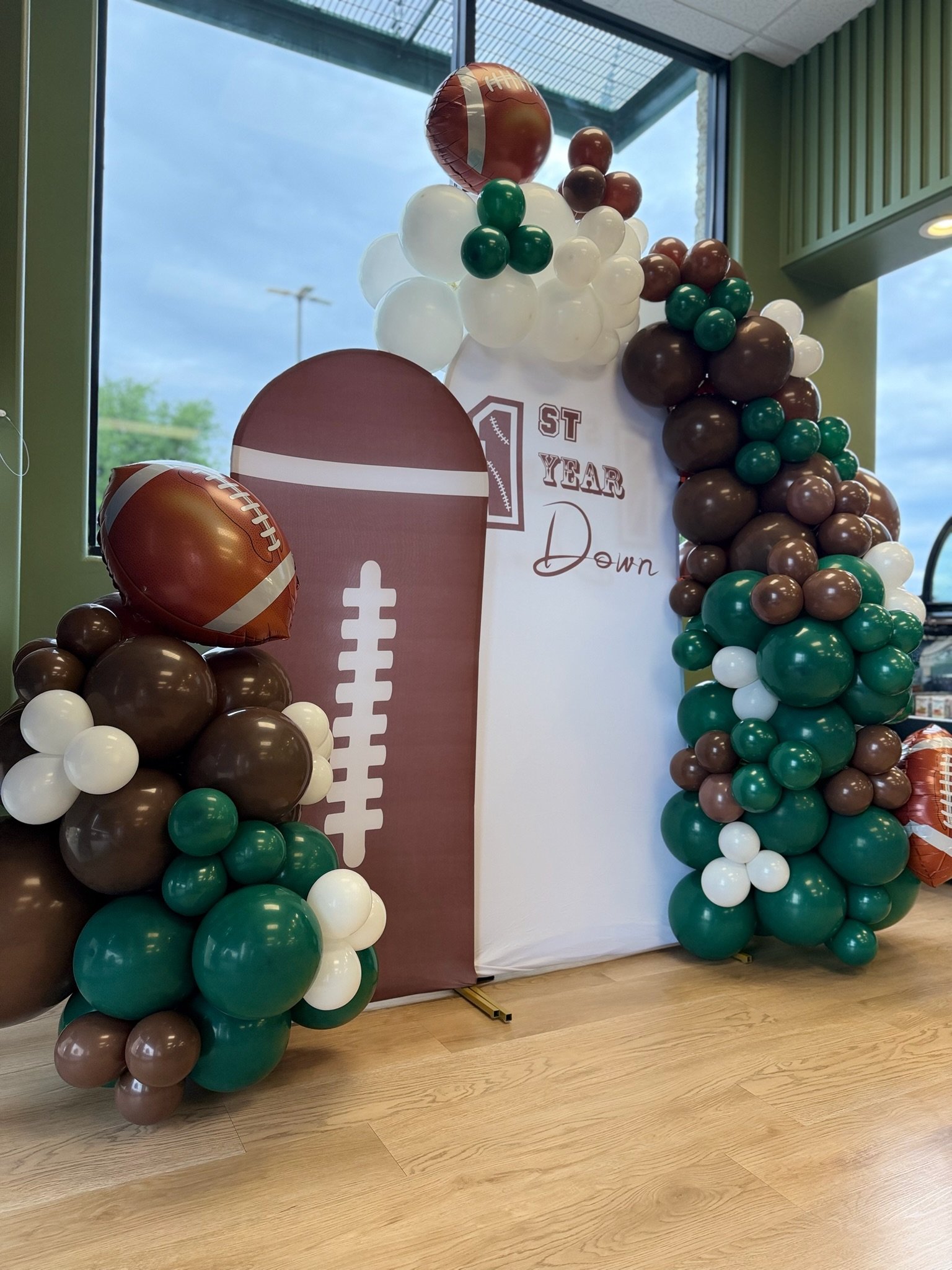 Indoor decoration featuring football-themed balloon arch with brown, white, and green balloons, and a sign celebrating a first year anniversary.
