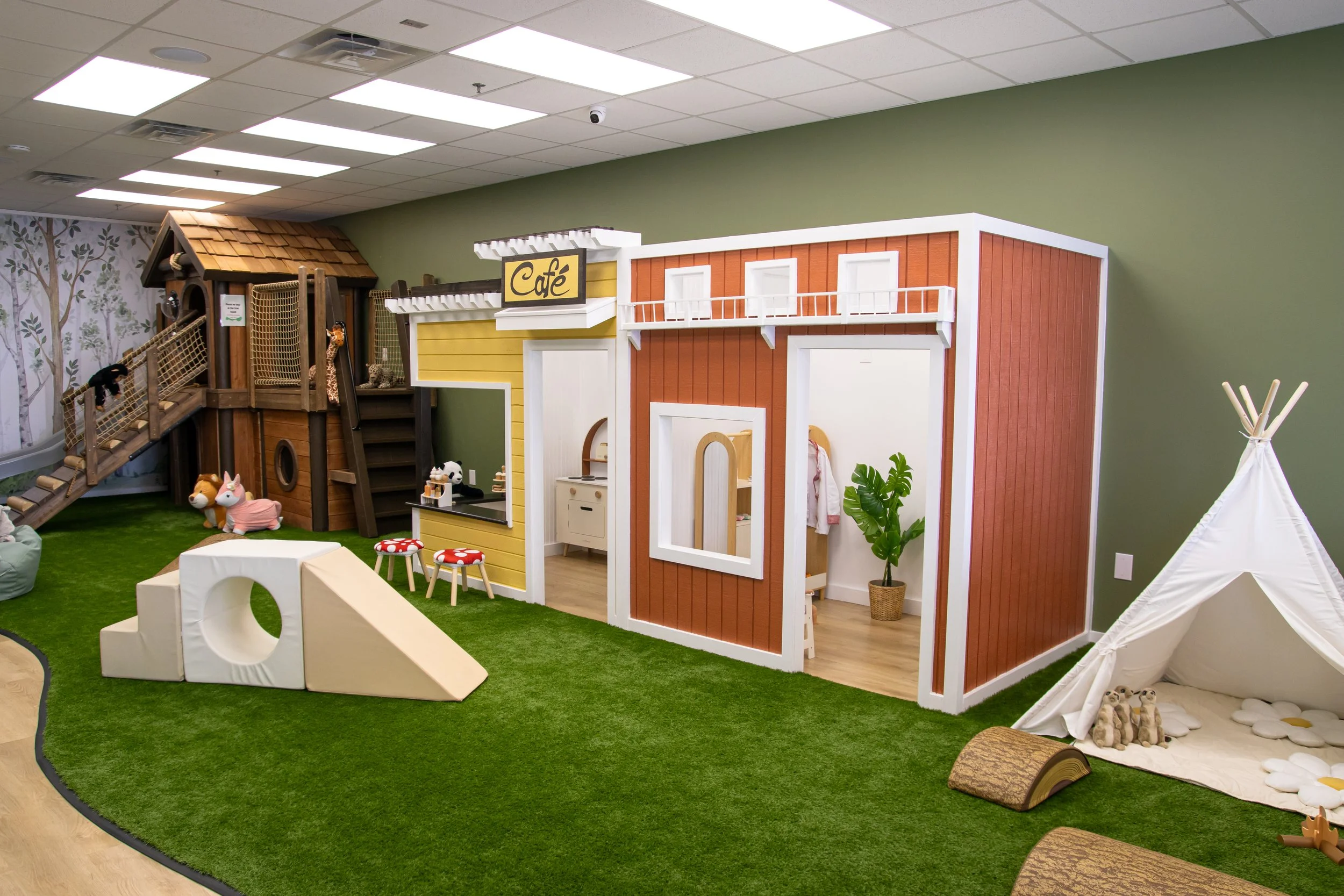 Indoor play area designed for children, featuring a wooden playhouse with a slide, plush toys on a green carpet, a teepee tent, and a small play kitchen with a sign that says 'Café,' decorated with plants and soft flooring.