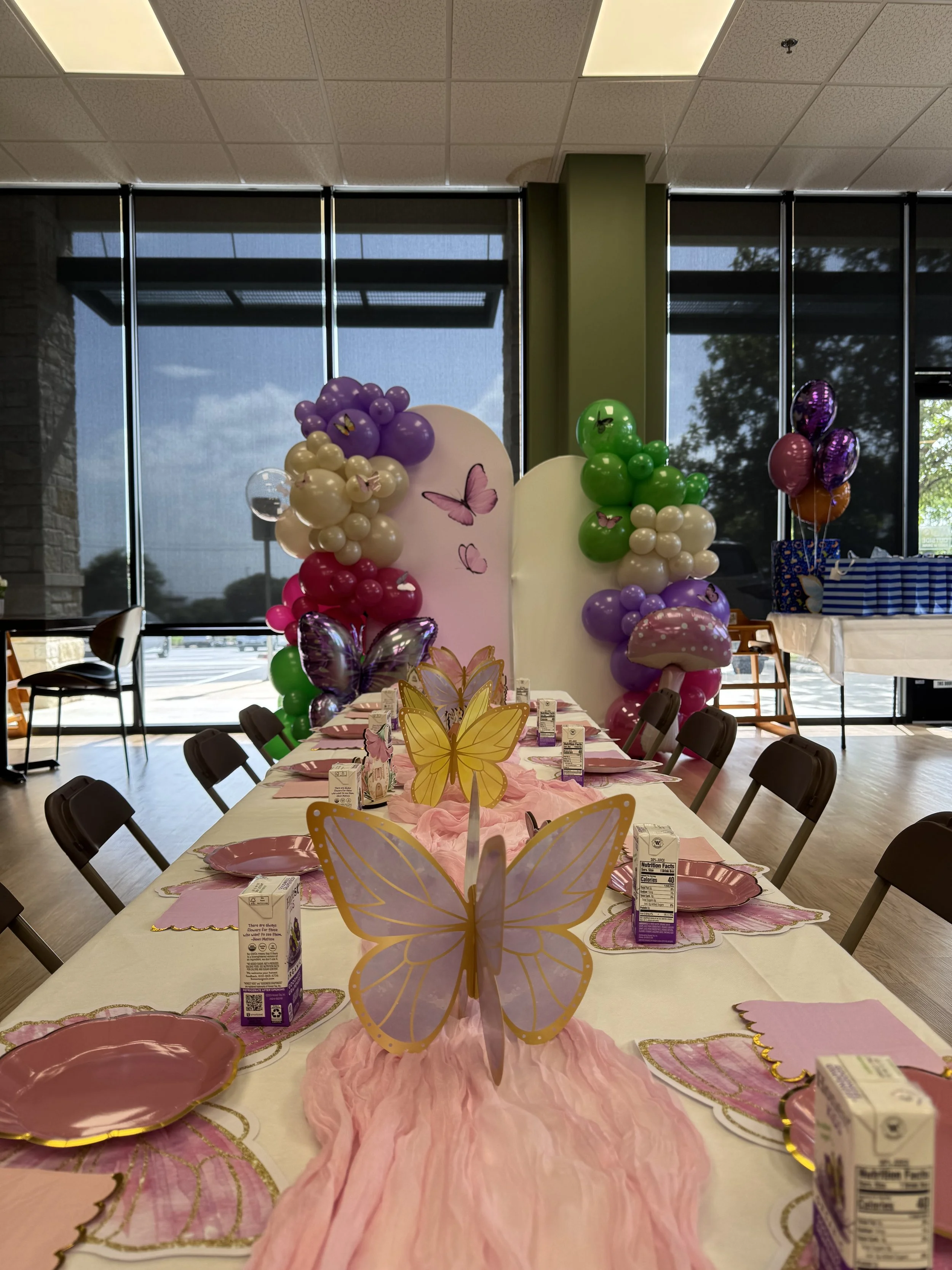 A decorated party table with pink and butterfly theme, featuring a pink table runner, butterfly centerpieces, pink paper plates, pink napkins, and juice boxes. In the background, there are balloons in purple, green, cream, and pink with butterfly des