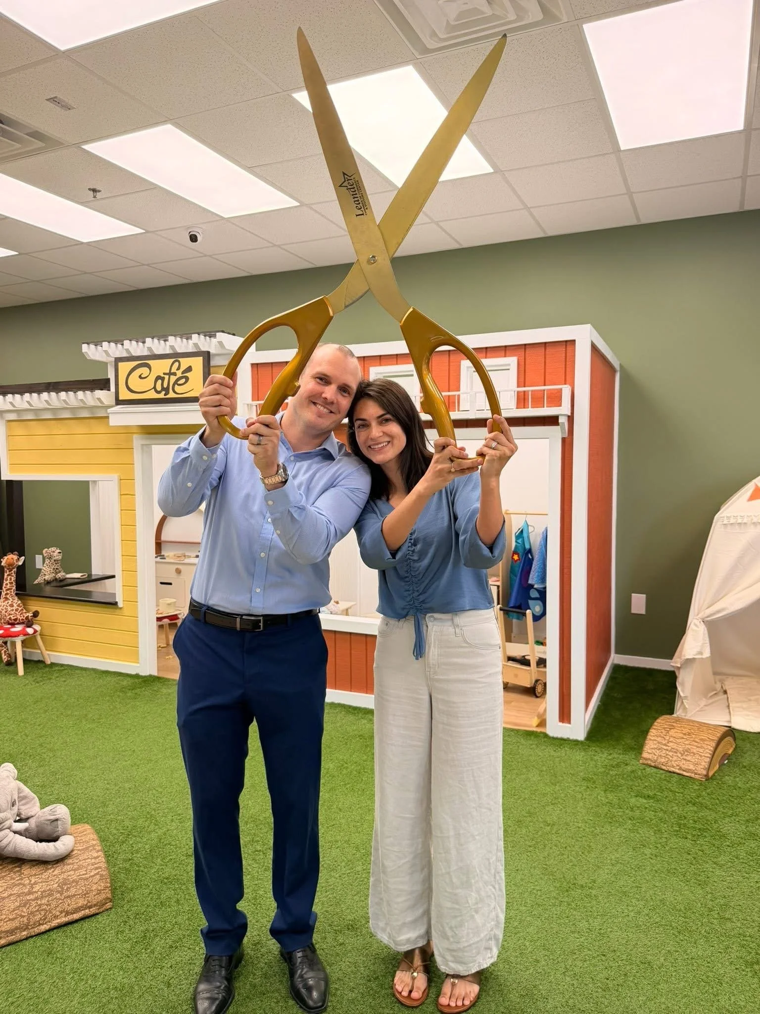 Two smiling people, a man and a woman, holding large yellow scissors together in an indoor setting with a children's play area visible in the background.
