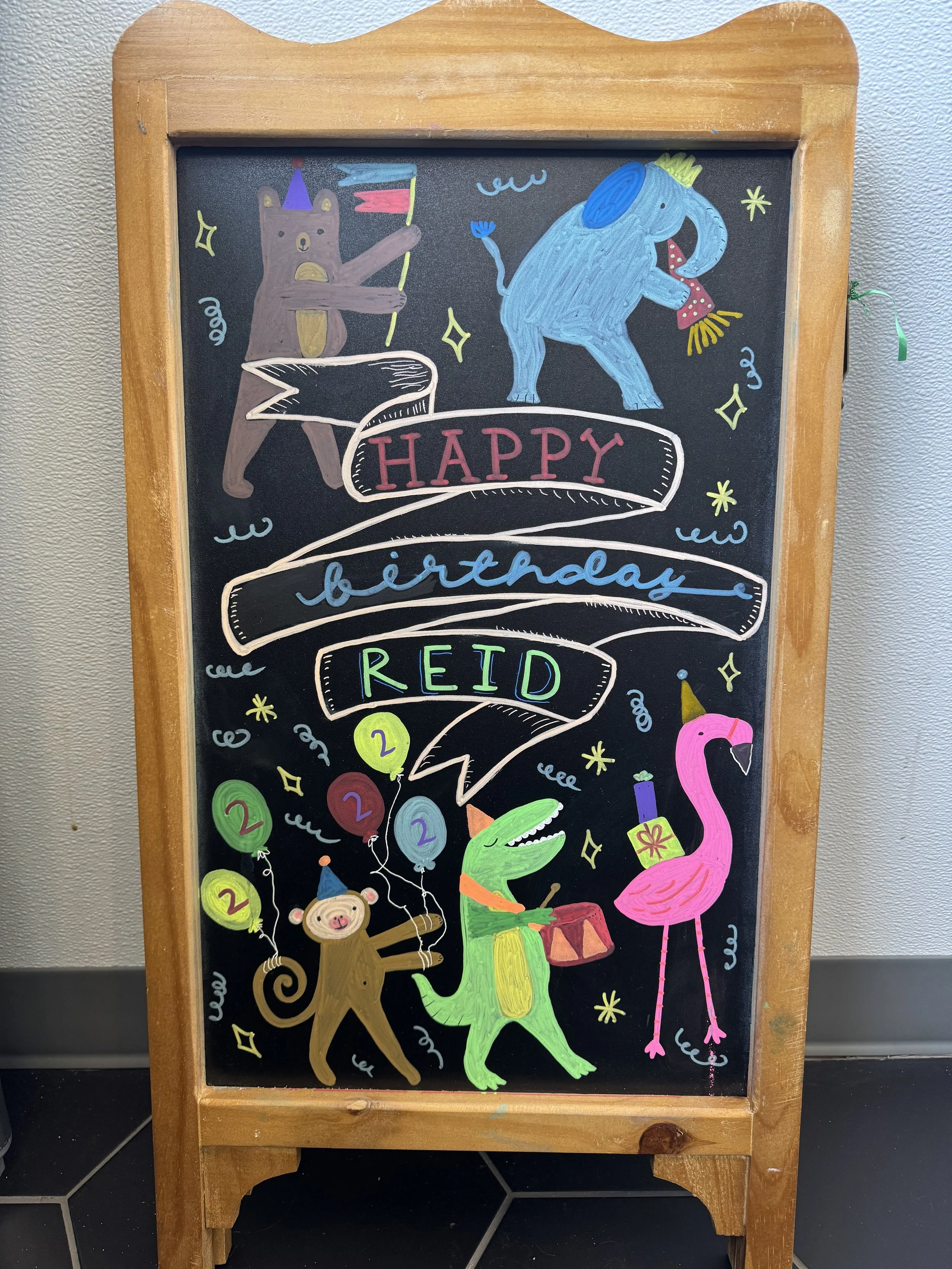 A colorful chalkboard drawing with animals celebrating a birthday. It features a bear wearing a party hat and holding a flag, a blue elephant wearing a party hat and blowing a party horn, a pink flamingo wearing a party hat, a monkey holding balloons